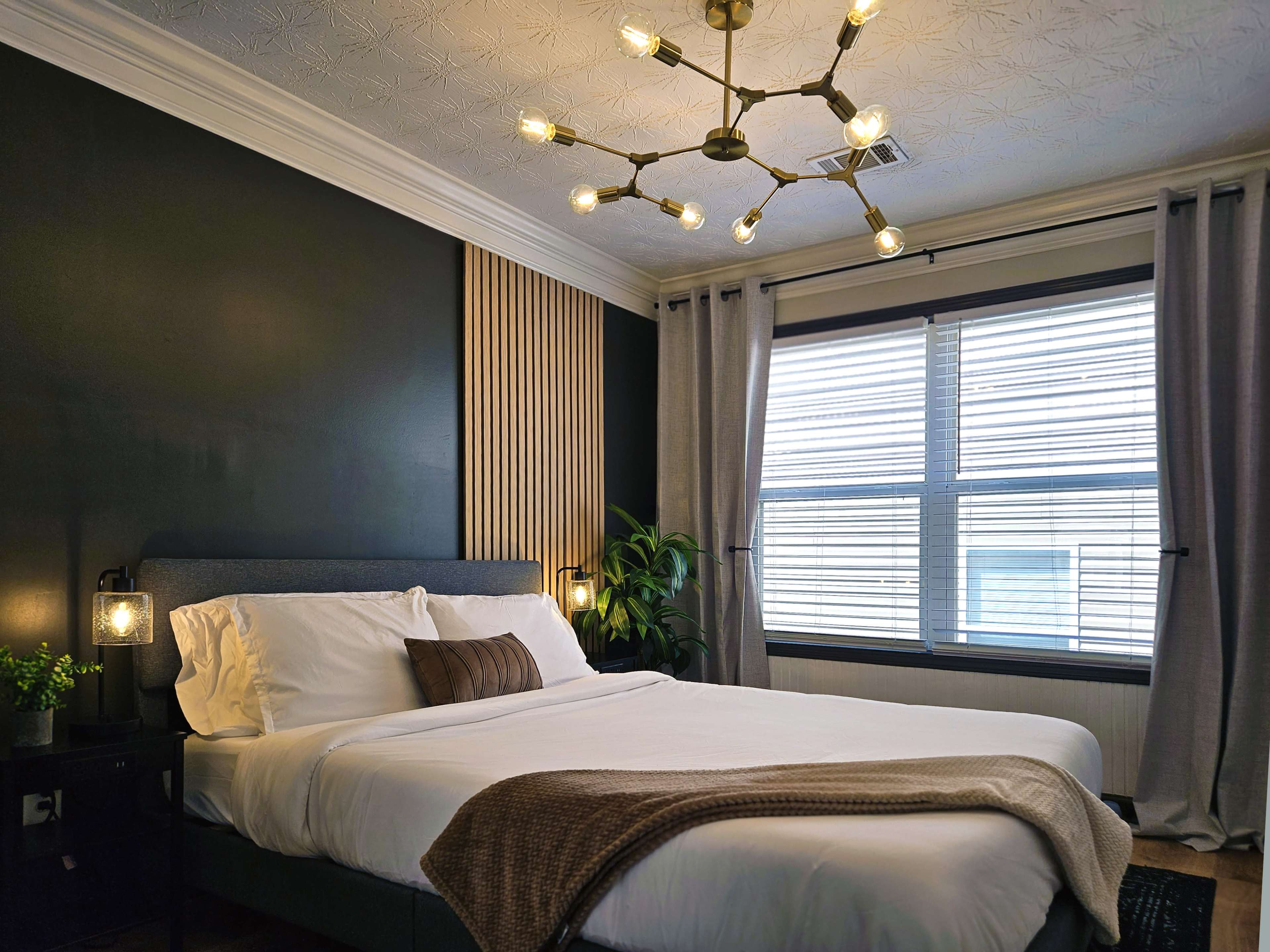 The image shows a modern bedroom featuring a gray bed with white linens, a decorative throw blanket, large windows with blinds, and a pendant light fixture.