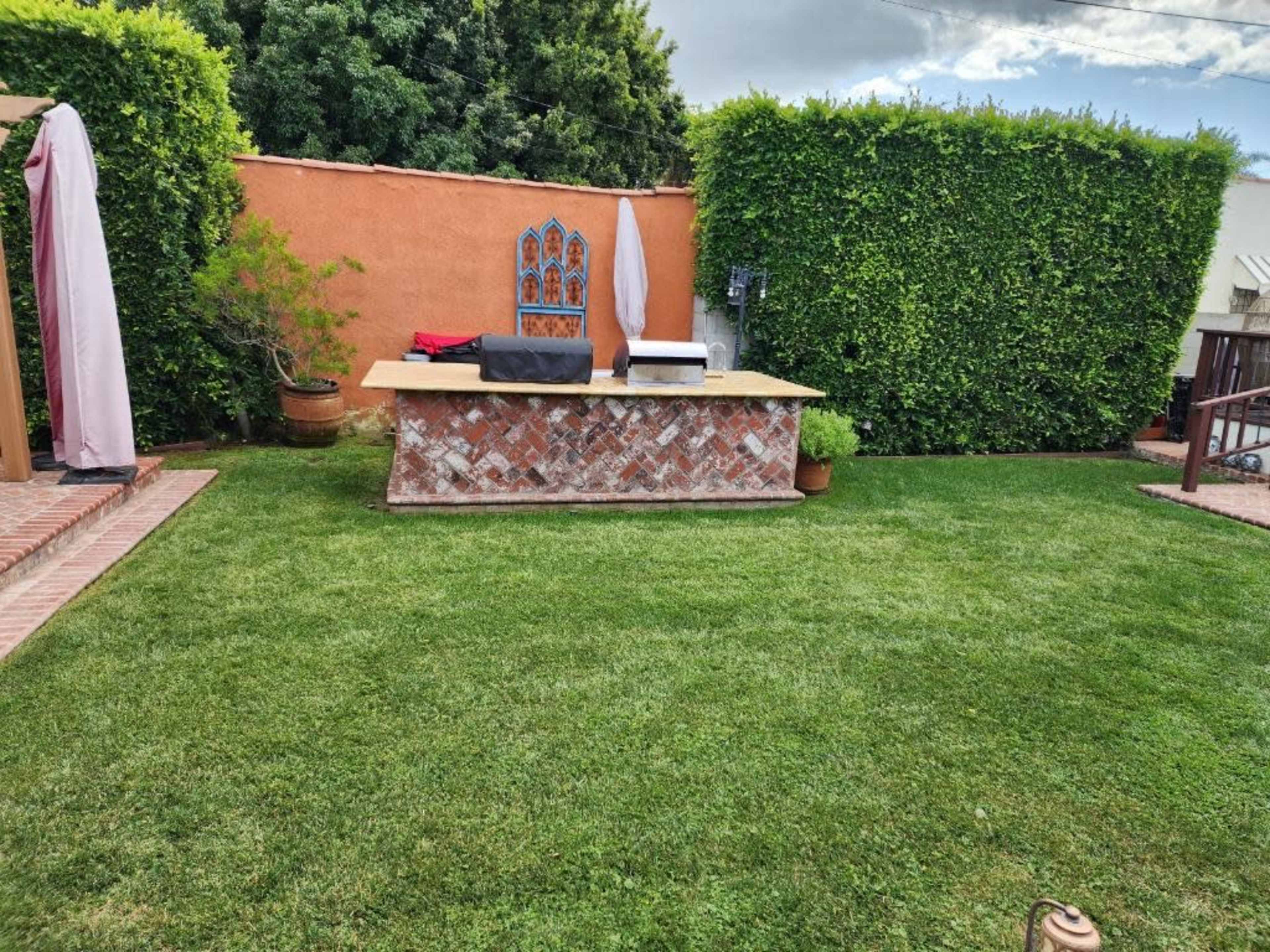 A well-maintained garden area featuring manicured grass, a stone barbecue island, and lush green hedges.