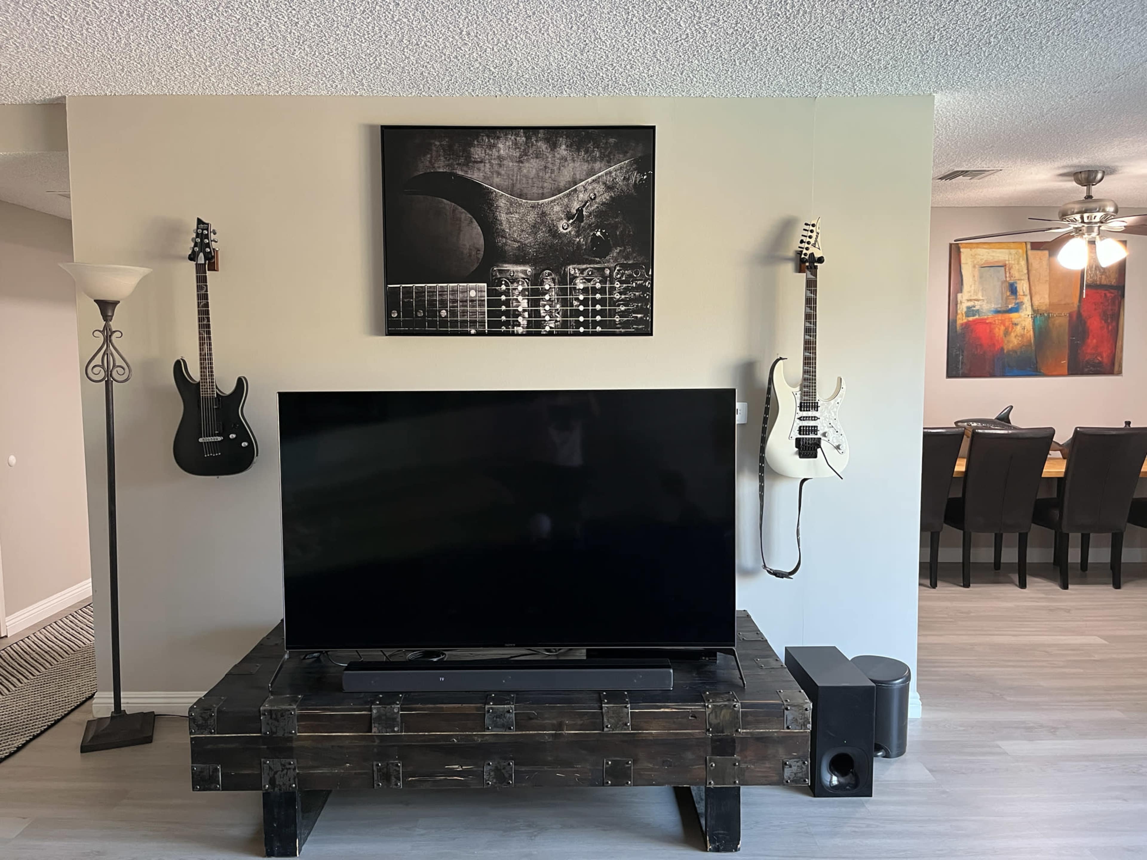 A living room with a large television on a rustic wooden stand, flanked by two electric guitars hanging on the wall above and a dining area visible in the background.