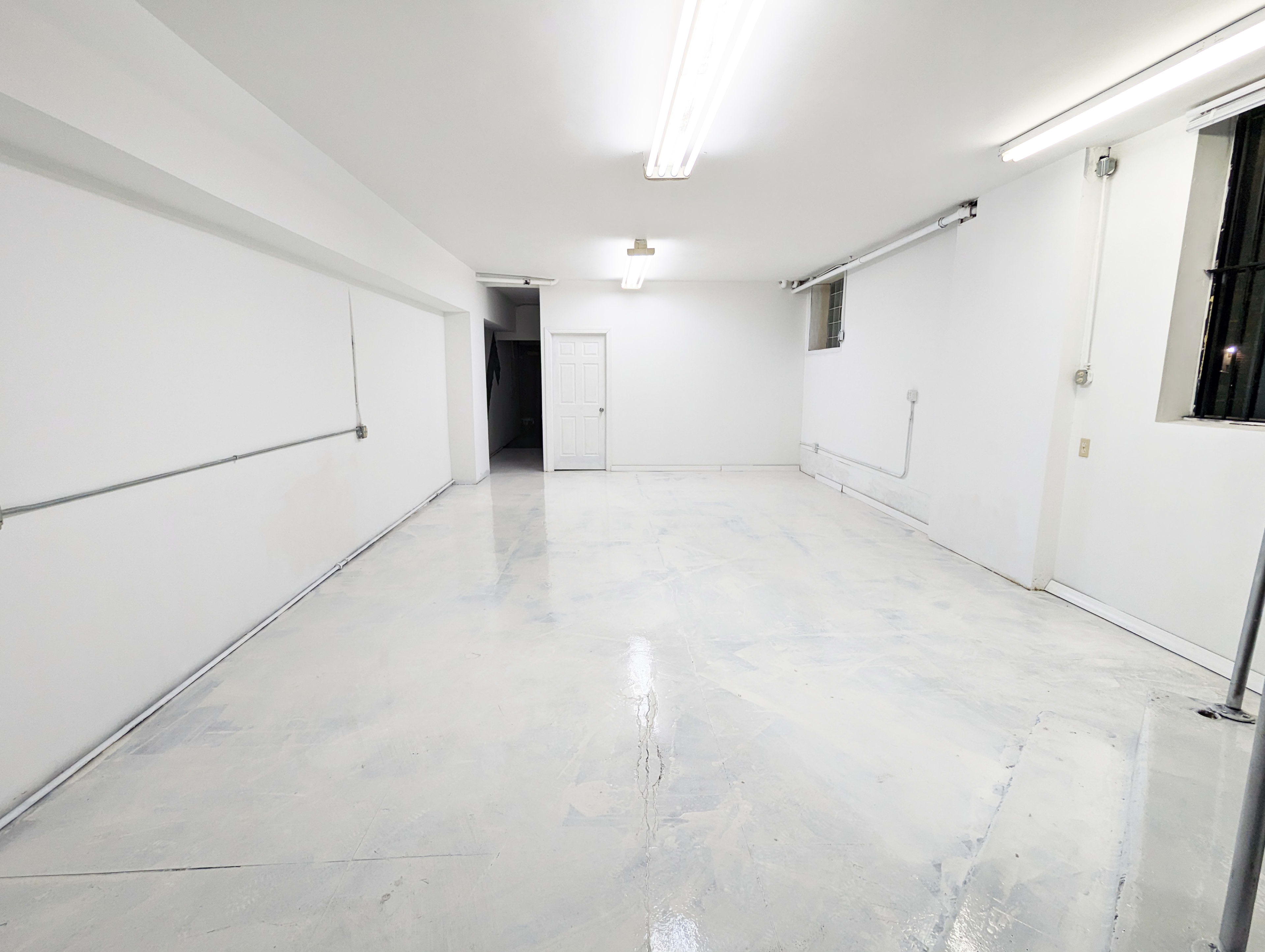 The image shows a large, empty room with white walls and a shiny, white-painted floor, illuminated by fluorescent lights.