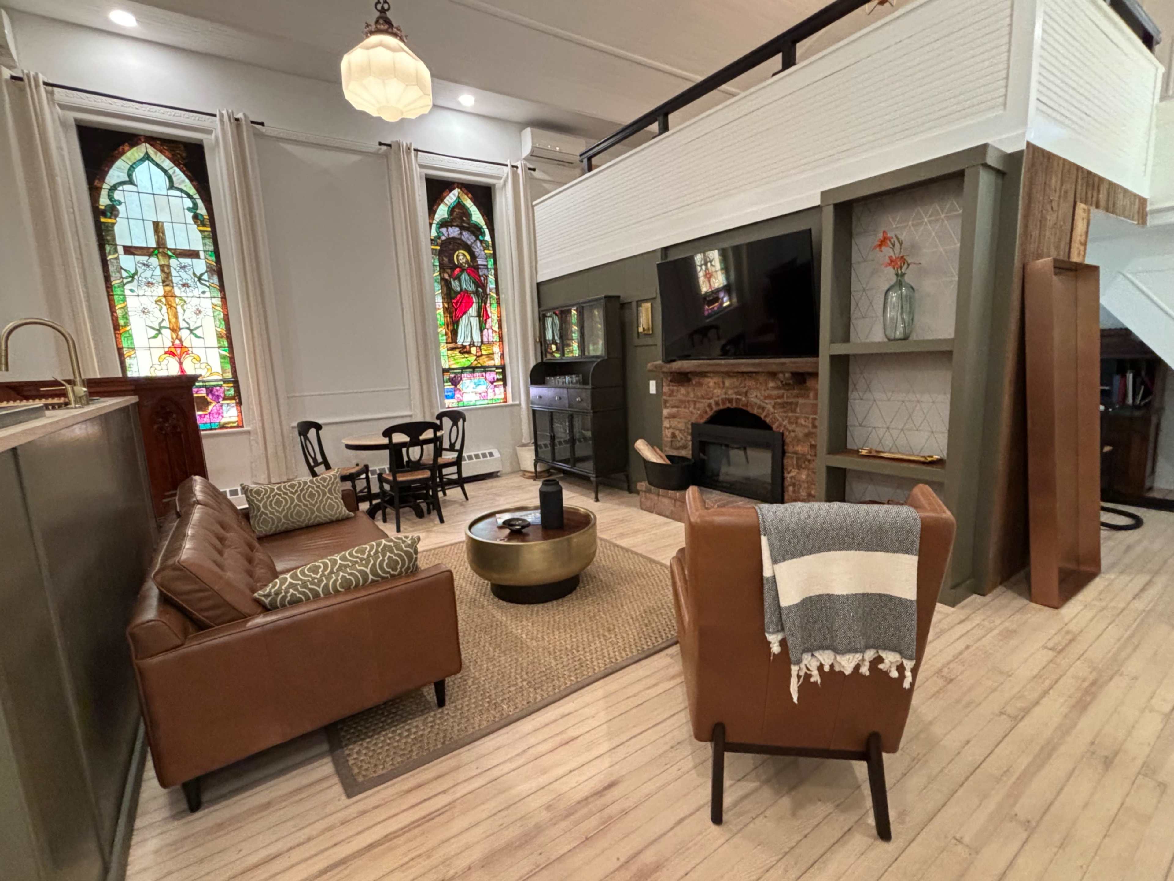A modern living room with a mix of contemporary furniture, a fireplace, and stained glass windows in a high-ceilinged space.