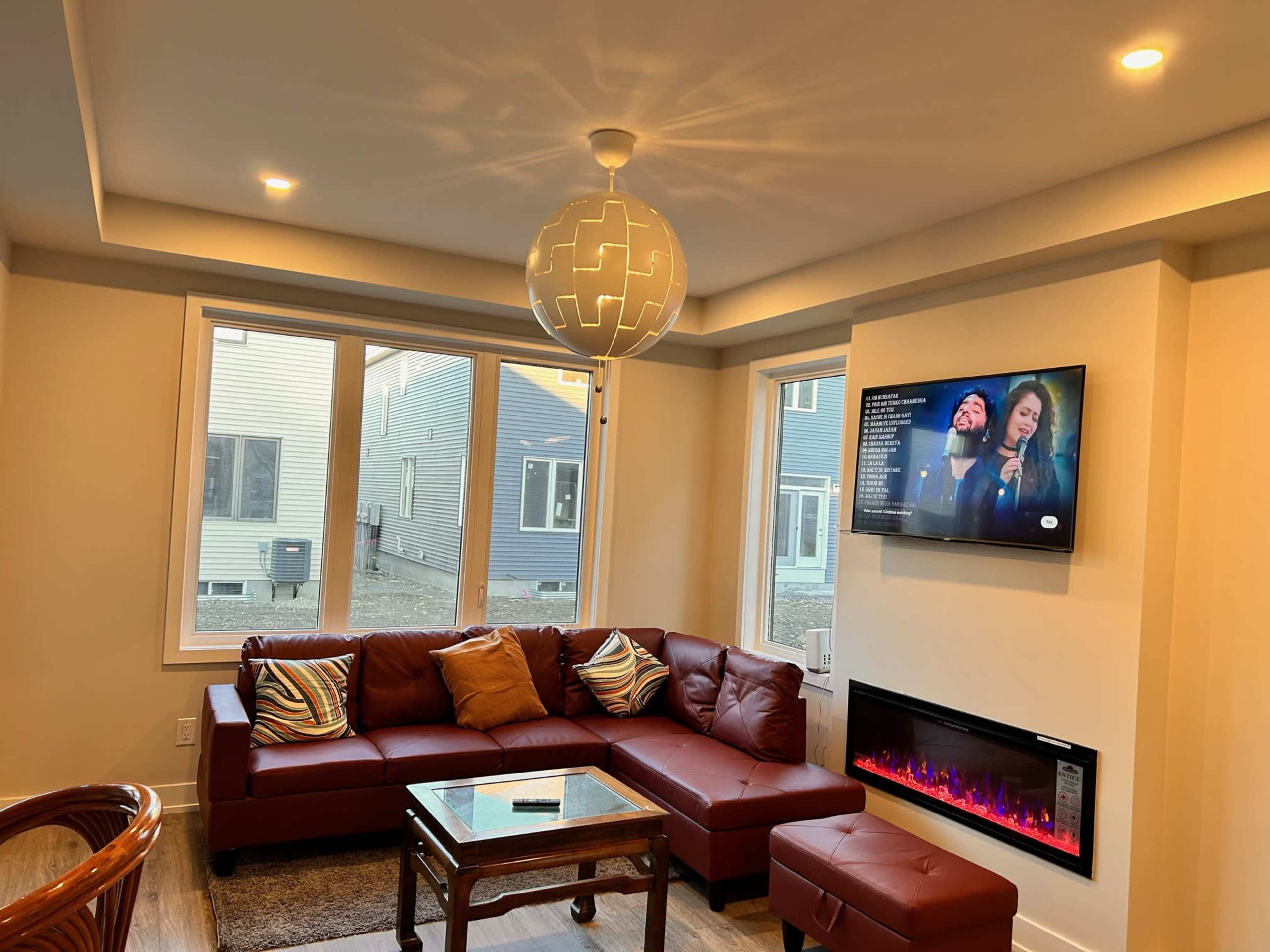 The image shows a modern living room featuring a brown sectional sofa, a wall-mounted television displaying a show, and a contemporary light fixture above a glass coffee table.