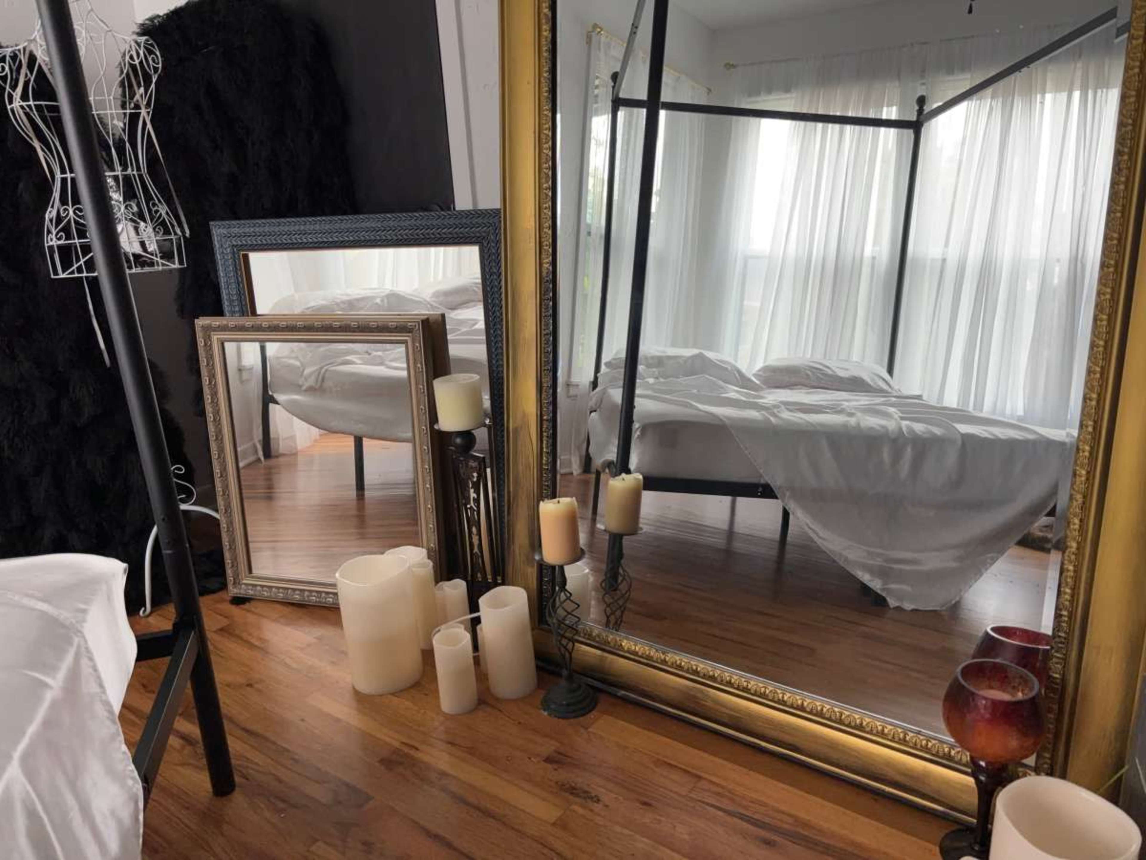 The image shows a bedroom scene featuring a four-poster bed reflected in a large mirror, surrounded by several candles and decorative frames on a wooden floor.