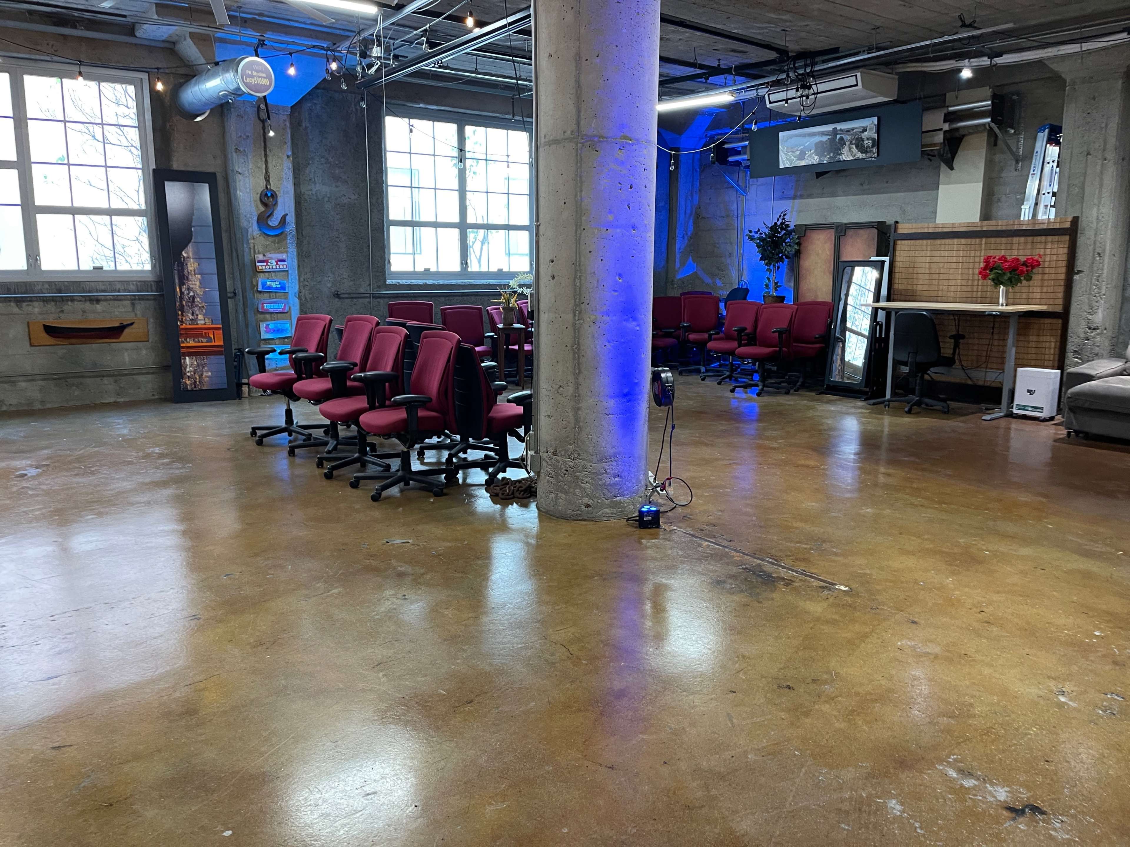 An interior space with several red office chairs arranged in rows, a concrete floor, and large windows allowing natural light.