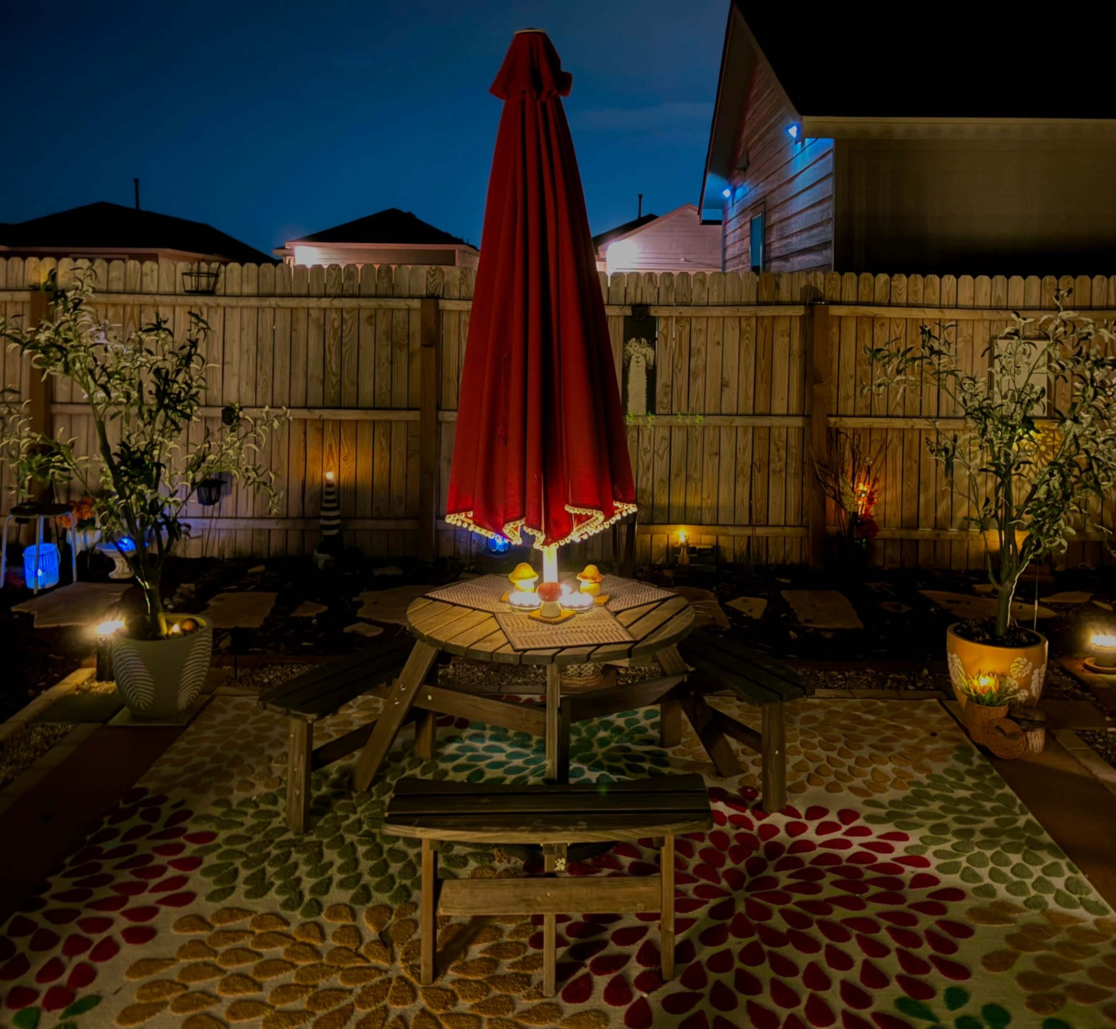 The image shows a backyard scene at night featuring a round table with a red umbrella, surrounded by wooden benches, colorful rugs, and illuminated plants along a wooden fence.