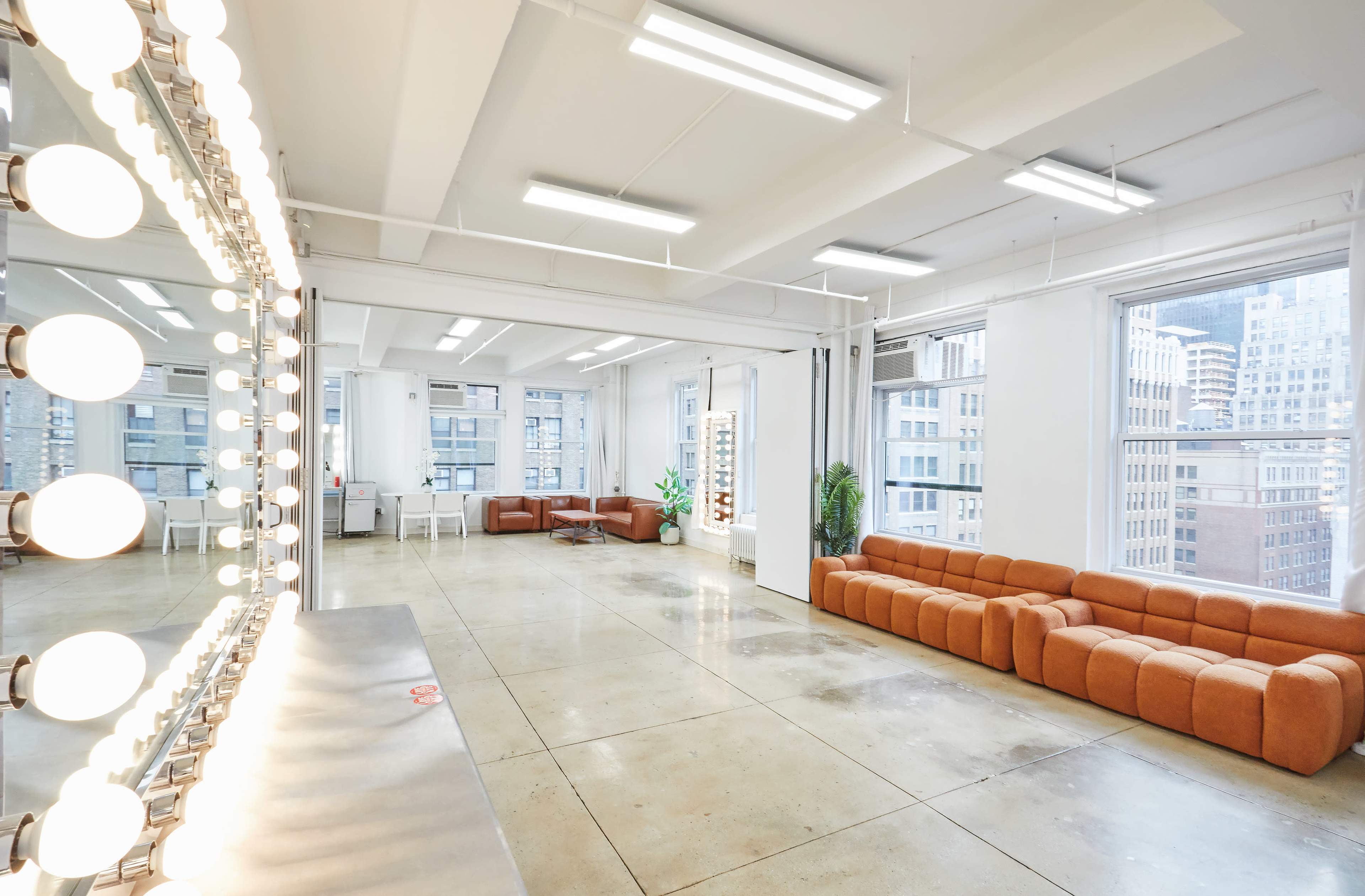 The image shows a bright, spacious room with large windows, featuring a long sectional couch, a vanity with lights, and a polished concrete floor.