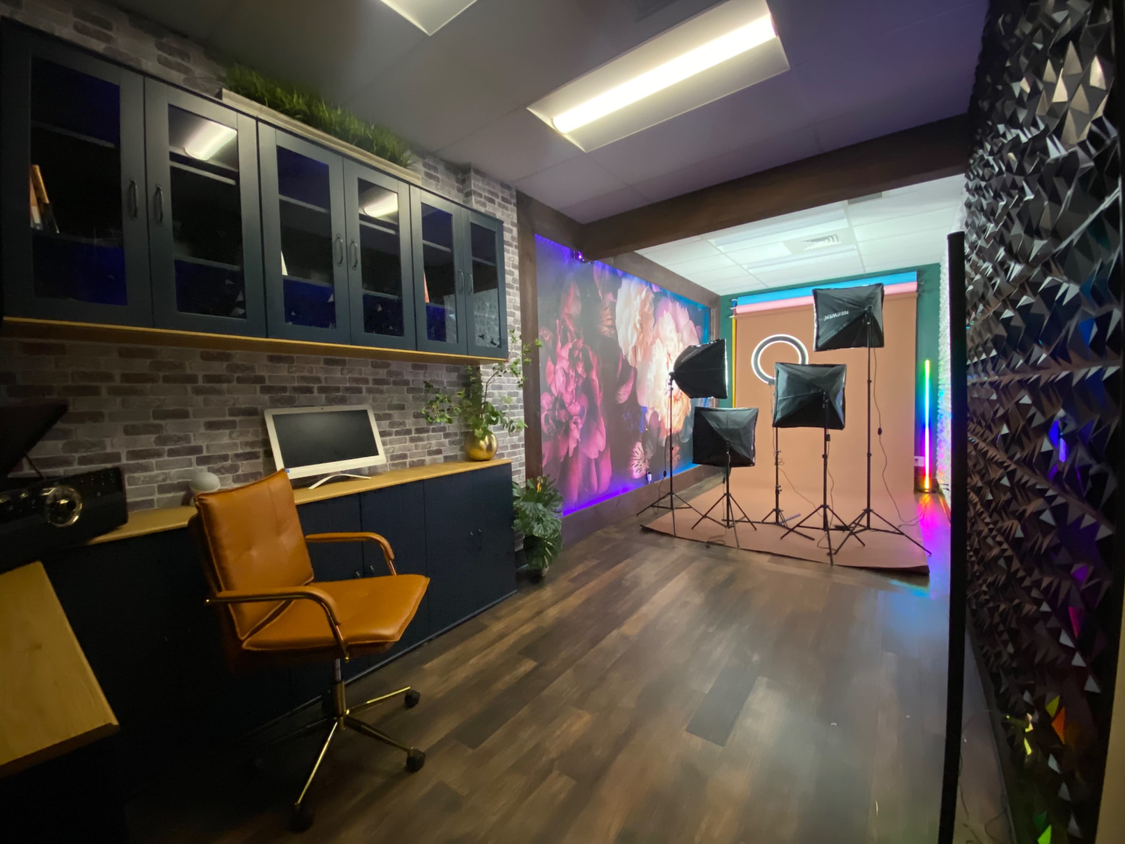 The image shows a photo studio featuring a wooden floor, a desk with an orange chair, and multiple lighting setups in front of a colorful backdrop.