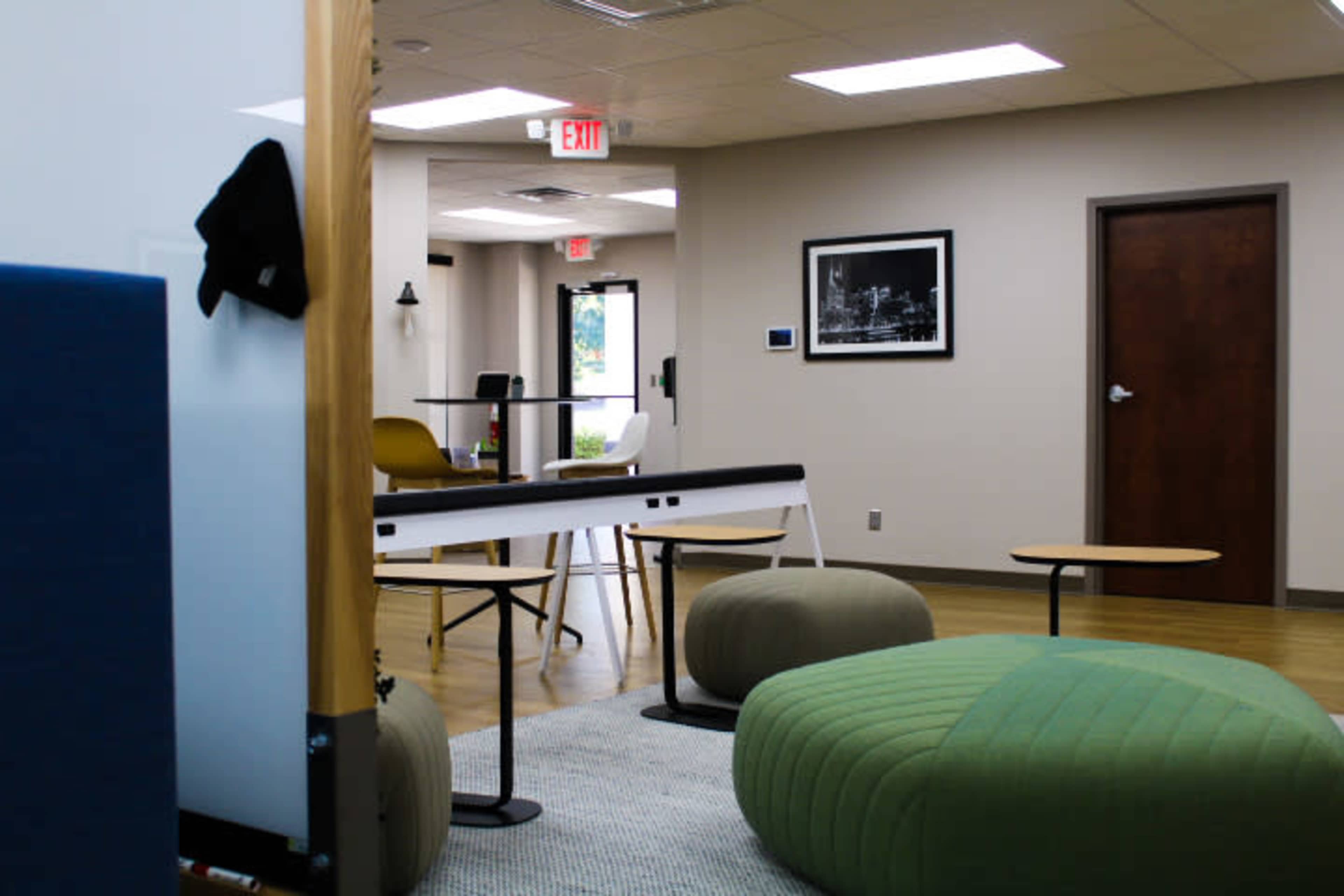 A modern office space featuring a seating area with round poufs, a coffee table, and chairs, with a hallway leading to an exit.