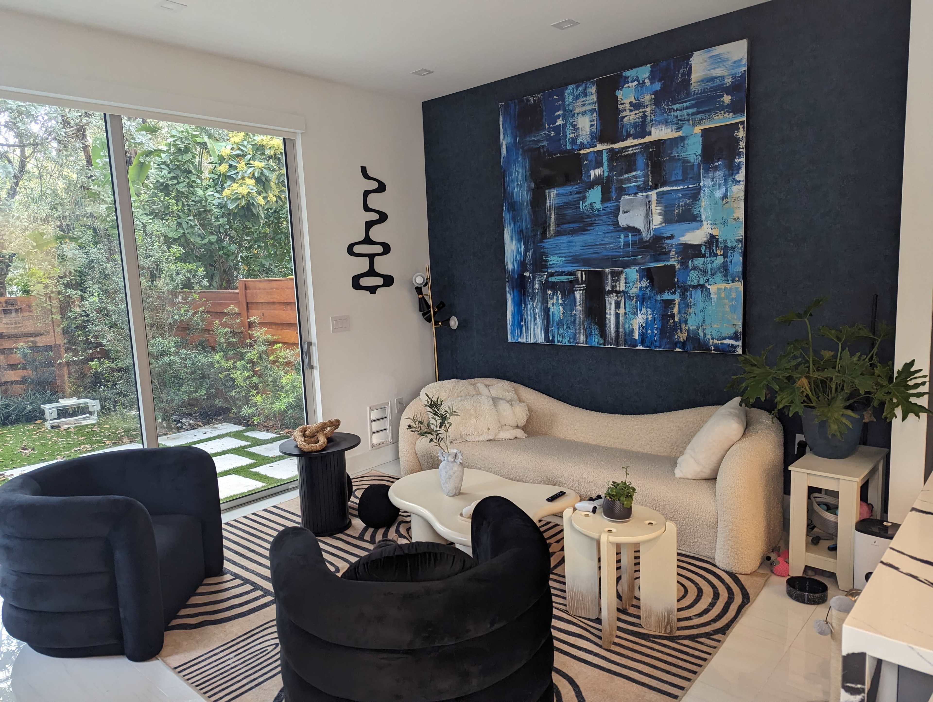 A modern living room features a curved white sofa, two black chairs, a coffee table, and a large blue abstract painting on the wall, with sliding glass doors opening to a garden.