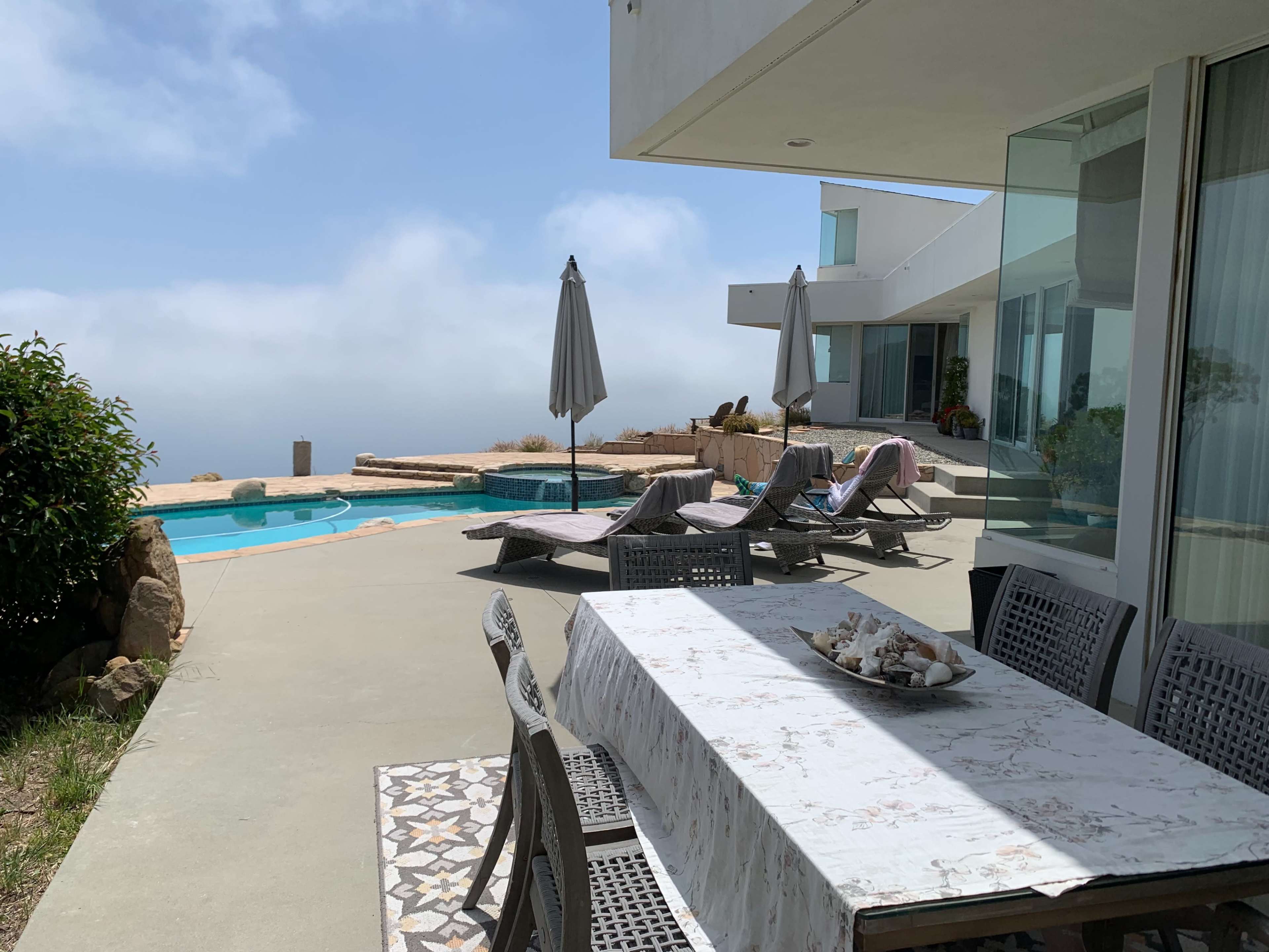 A patio area features a dining table and lounge chairs near a swimming pool, with partially cloudy skies in the background.