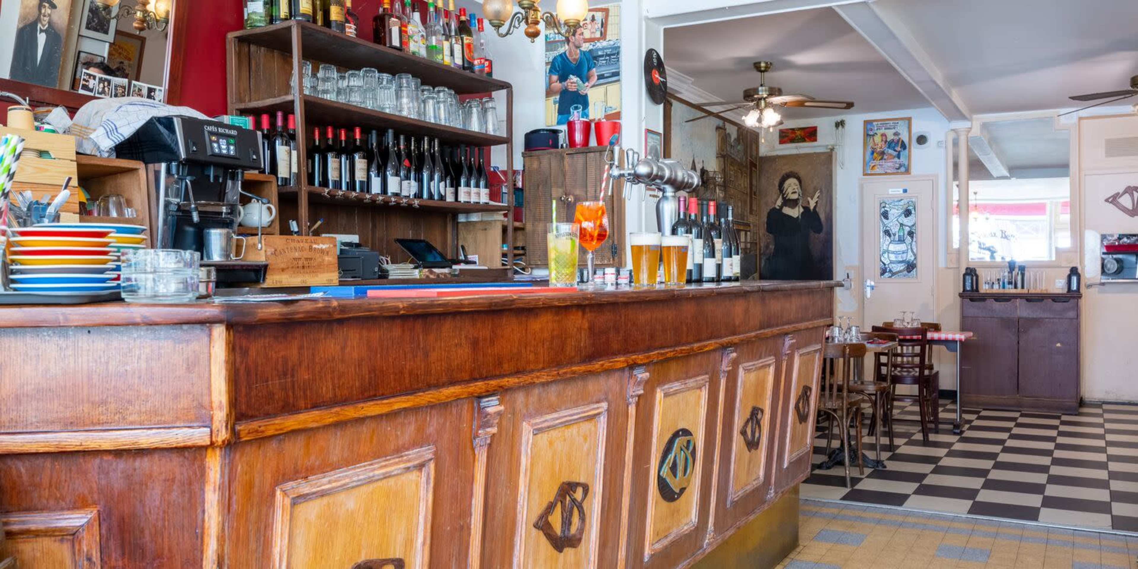 The image shows a cozy bar area with a wooden counter, shelves stocked with bottles, and colorful plates stacked on the surface.