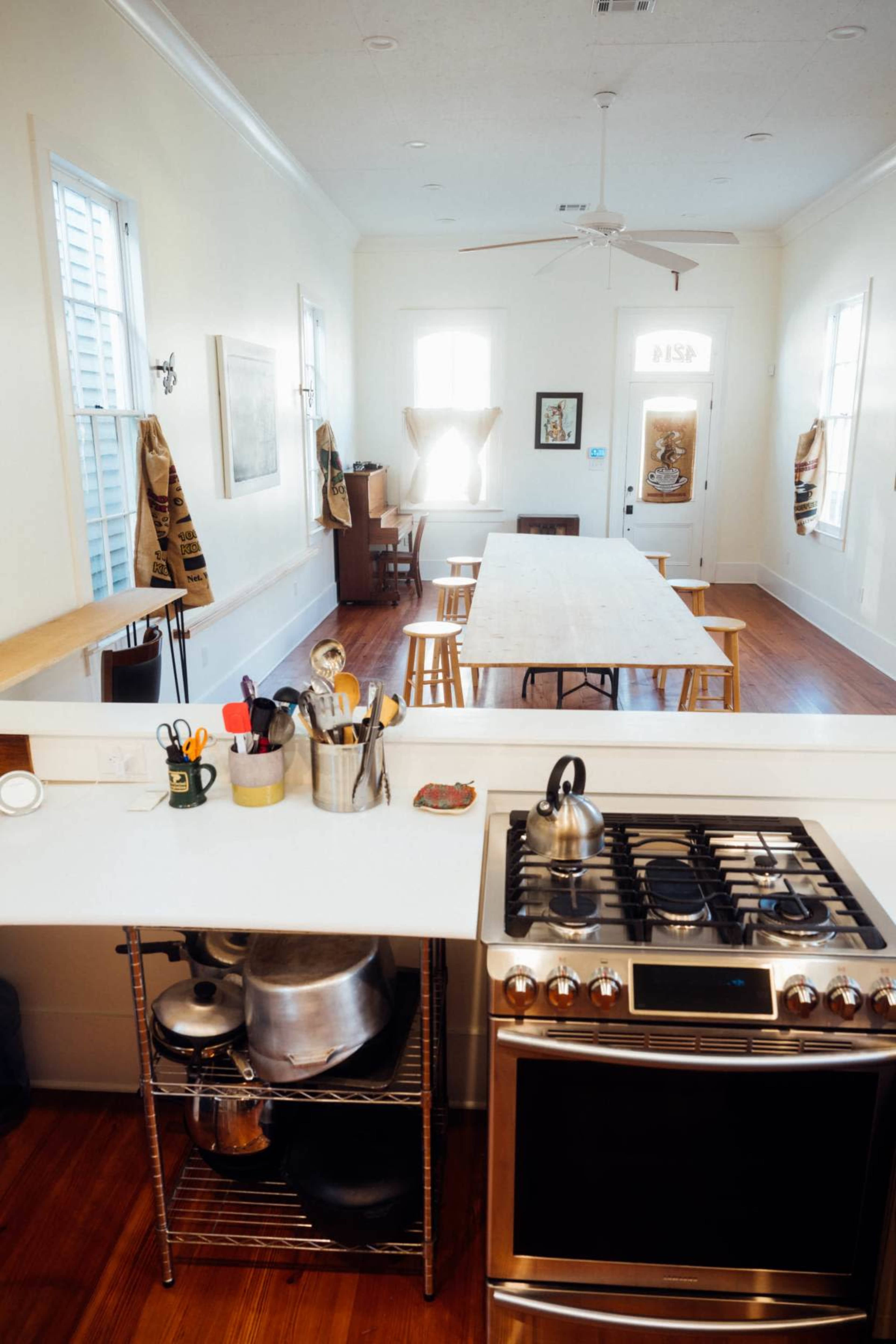 A kitchen with a stove, pots, and utensils in the foreground, leading to a large dining table and chairs in a bright, open space.
