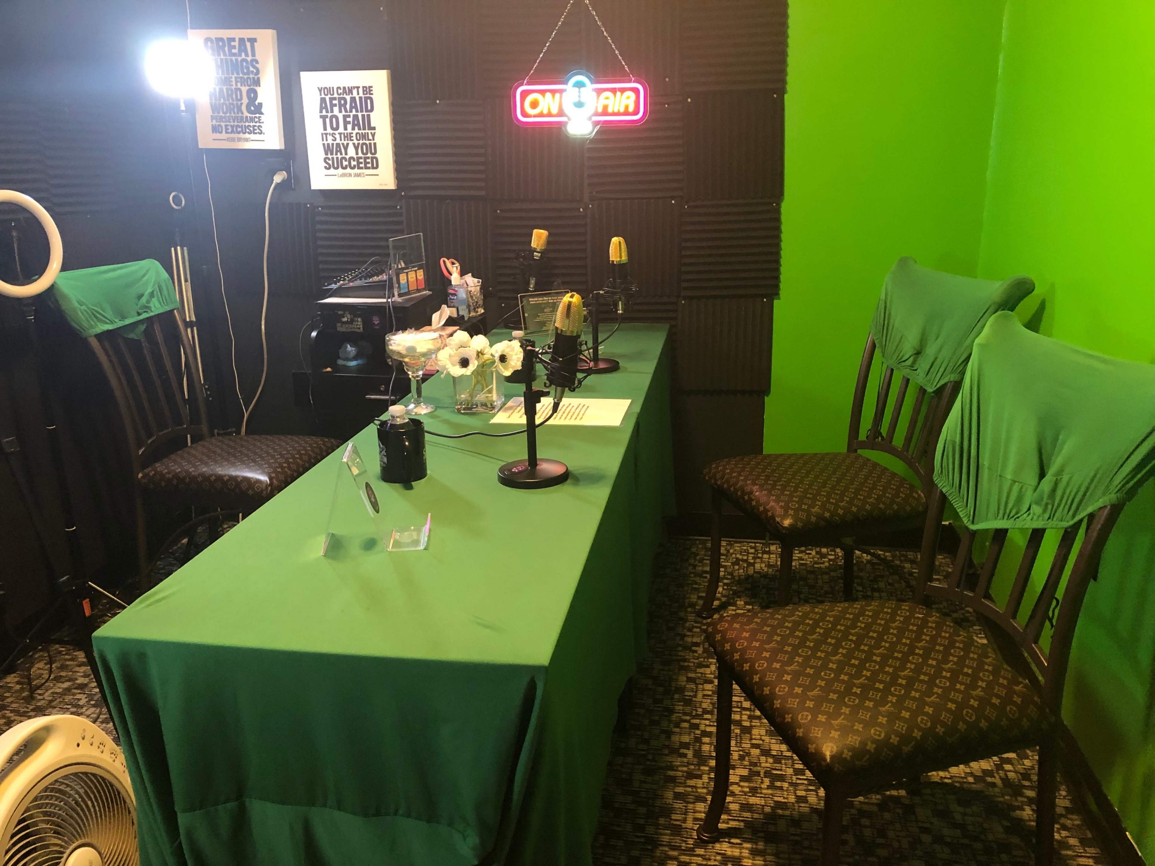 The image shows a recording studio with a green backdrop, a table covered in a green cloth, microphones, and chairs set up for a broadcast.