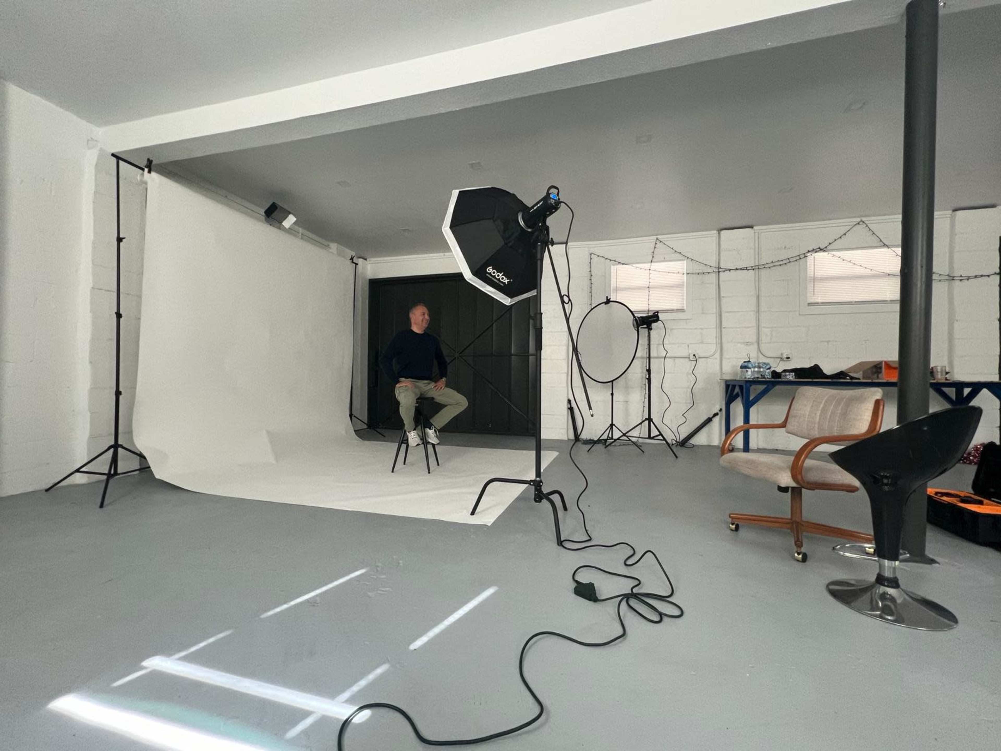 A person sits on a stool in a photo studio setup with a backdrop, lighting equipment, and props scattered around.