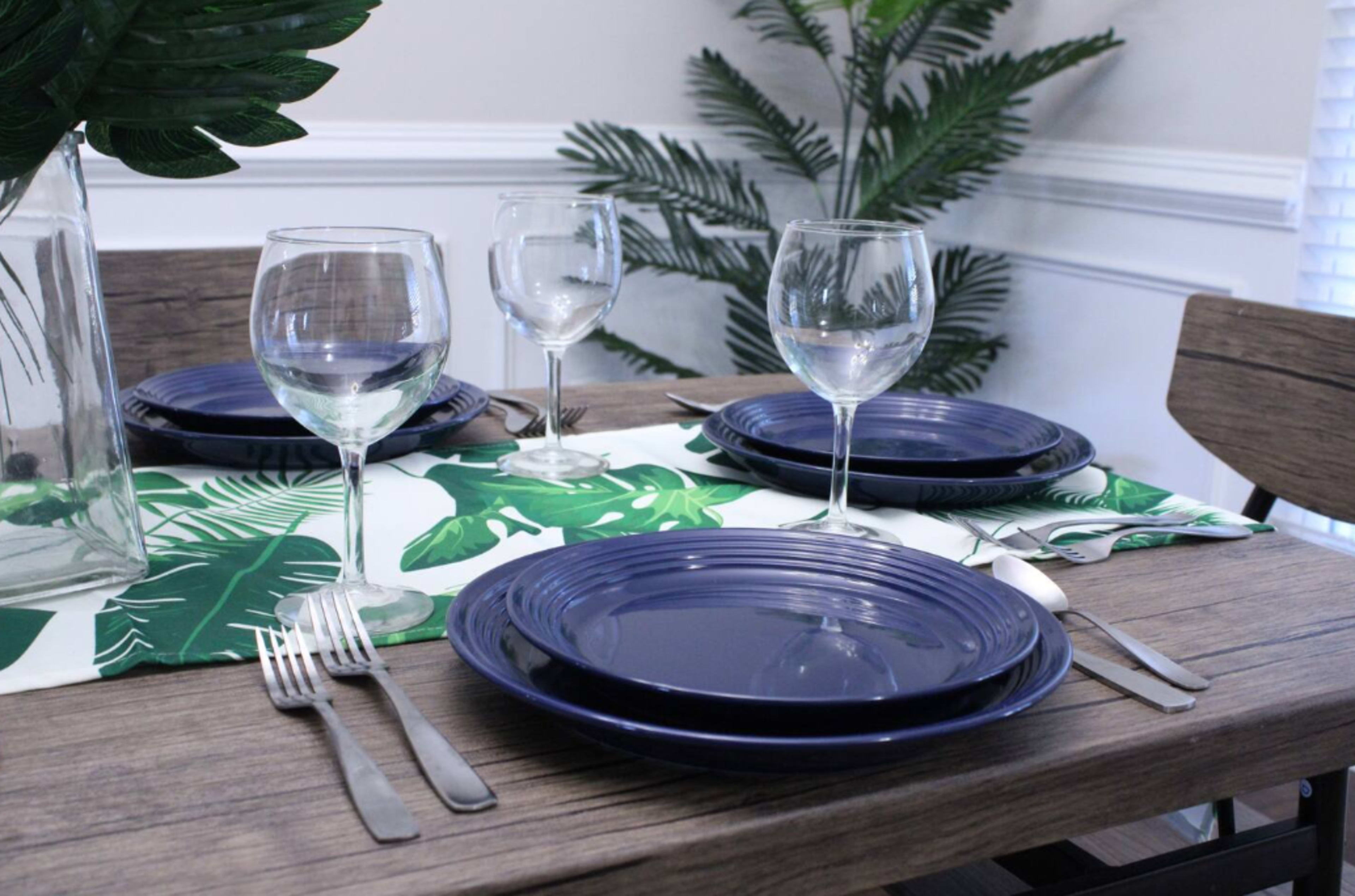 A dining table is set with navy plates, clear wine glasses, and silver cutlery, accompanied by a green leaf-patterned table runner and potted plants in the background.