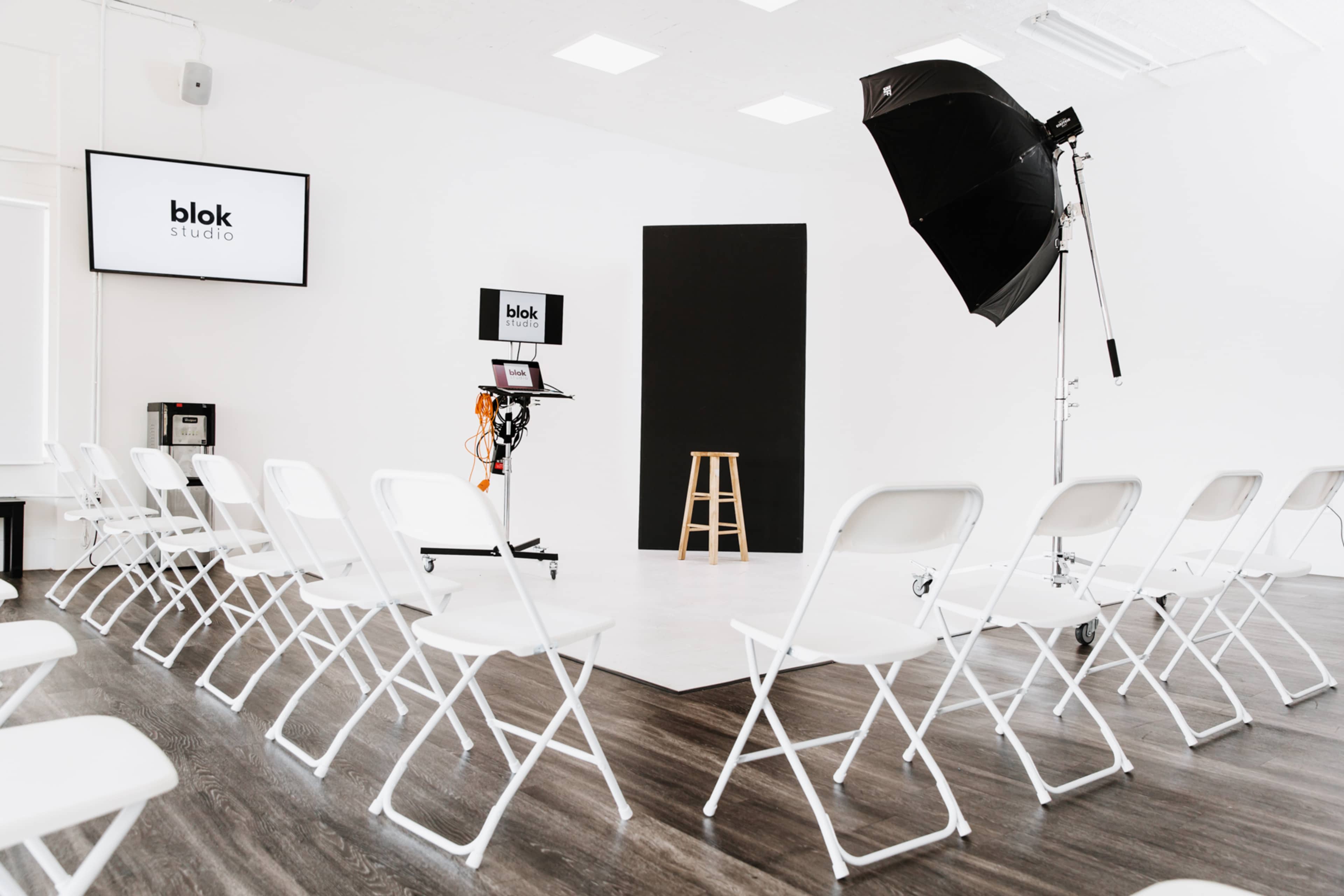 A minimalist studio setup with white chairs arranged in a semicircle around a small stage, featuring a black backdrop and lighting equipment.