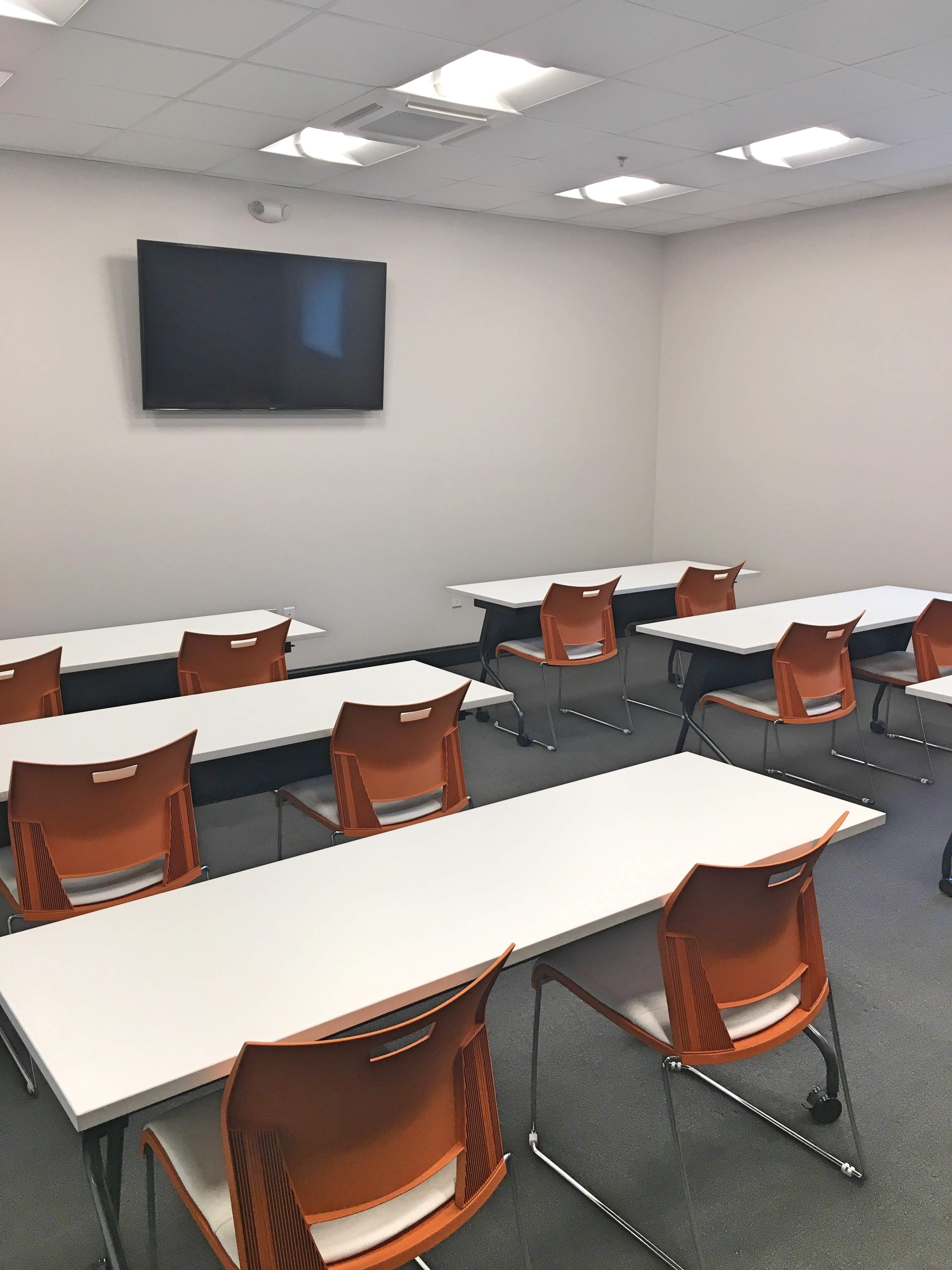 A modern classroom features several long white tables arranged with orange chairs and a wall-mounted television.