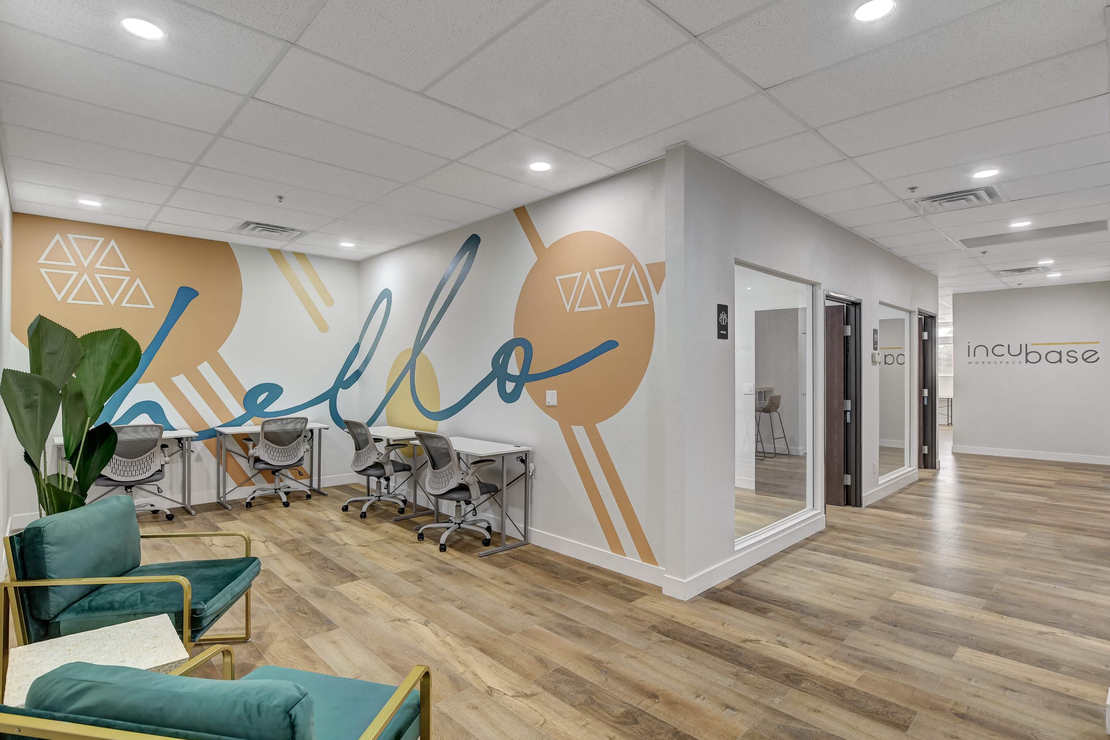 A modern office space featuring a large wall mural that says "Hello," a seating area with green chairs, and workstations along the walls.