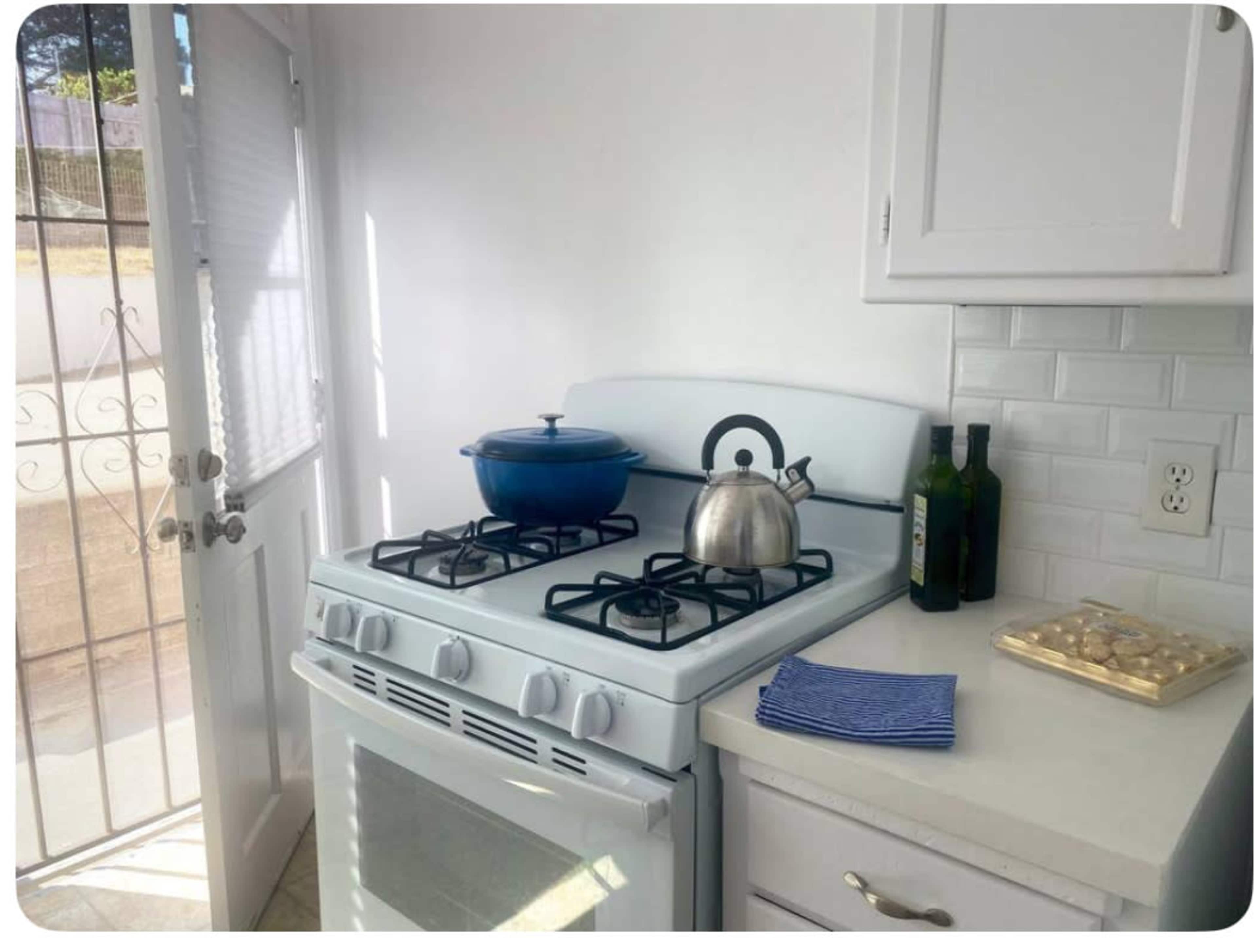 A white kitchen features a gas stove with a blue pot and a kettle on top, beside a window and a countertop with bottles and dishware.