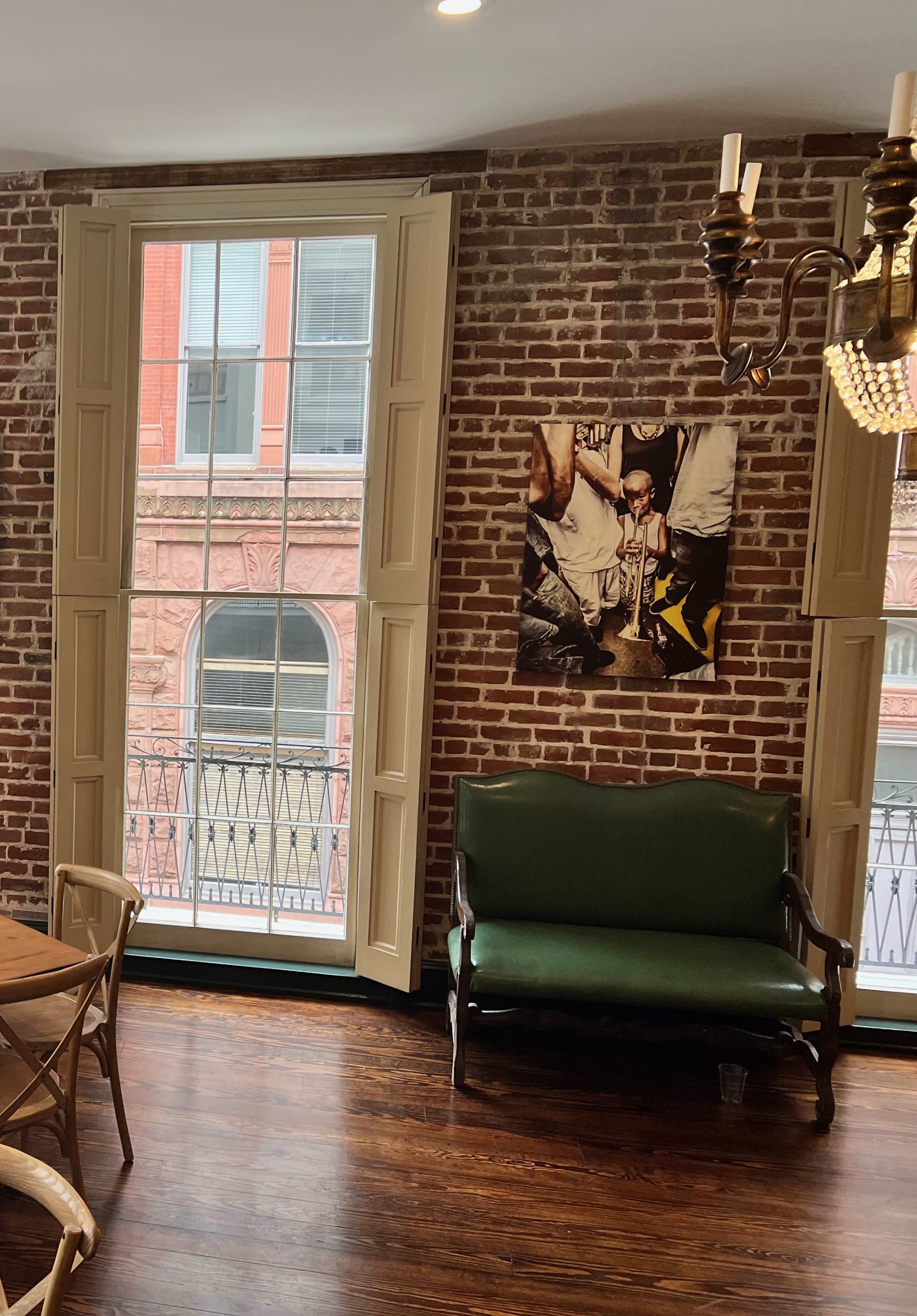 The scene shows a room with exposed brick walls, a green bench, and large windows revealing a view of a building's red façade outside.