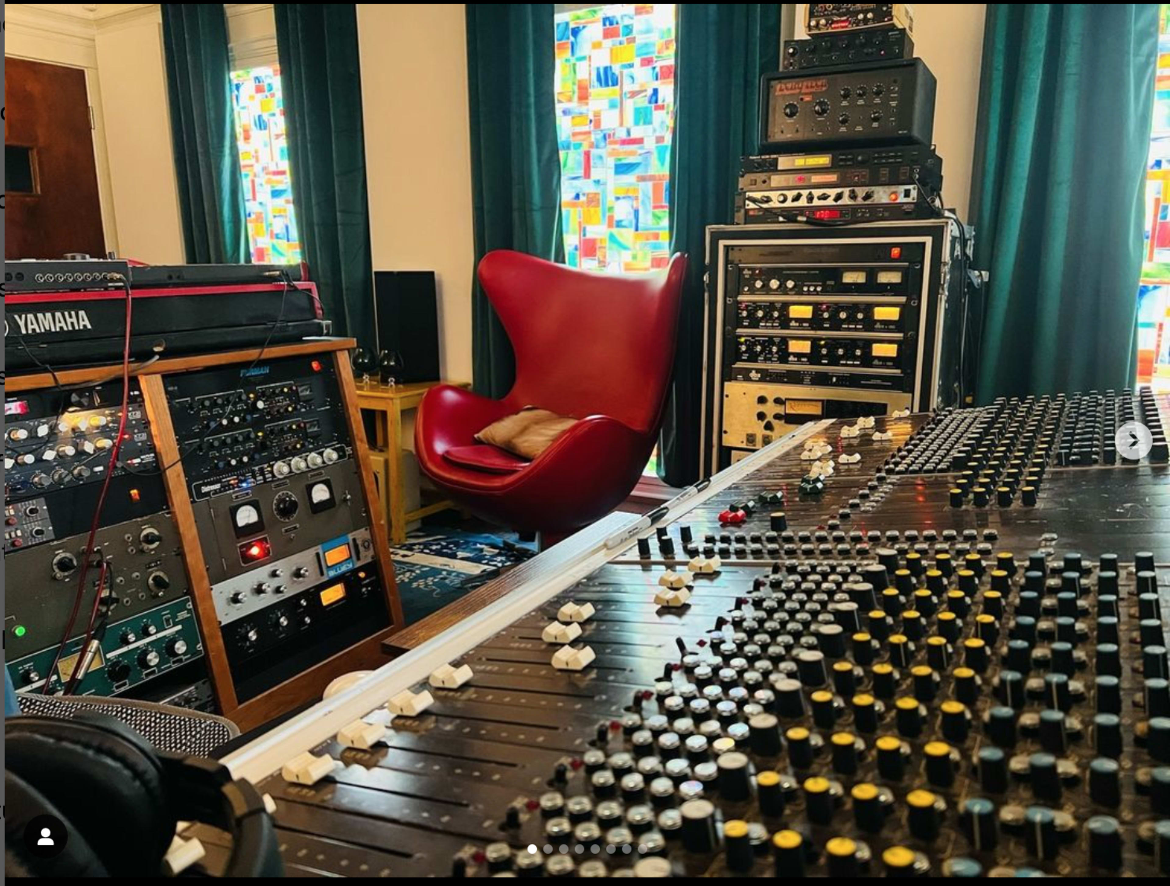 A music production studio features a large mixing console, various audio equipment racks, and a red chair positioned near colorful window panels.