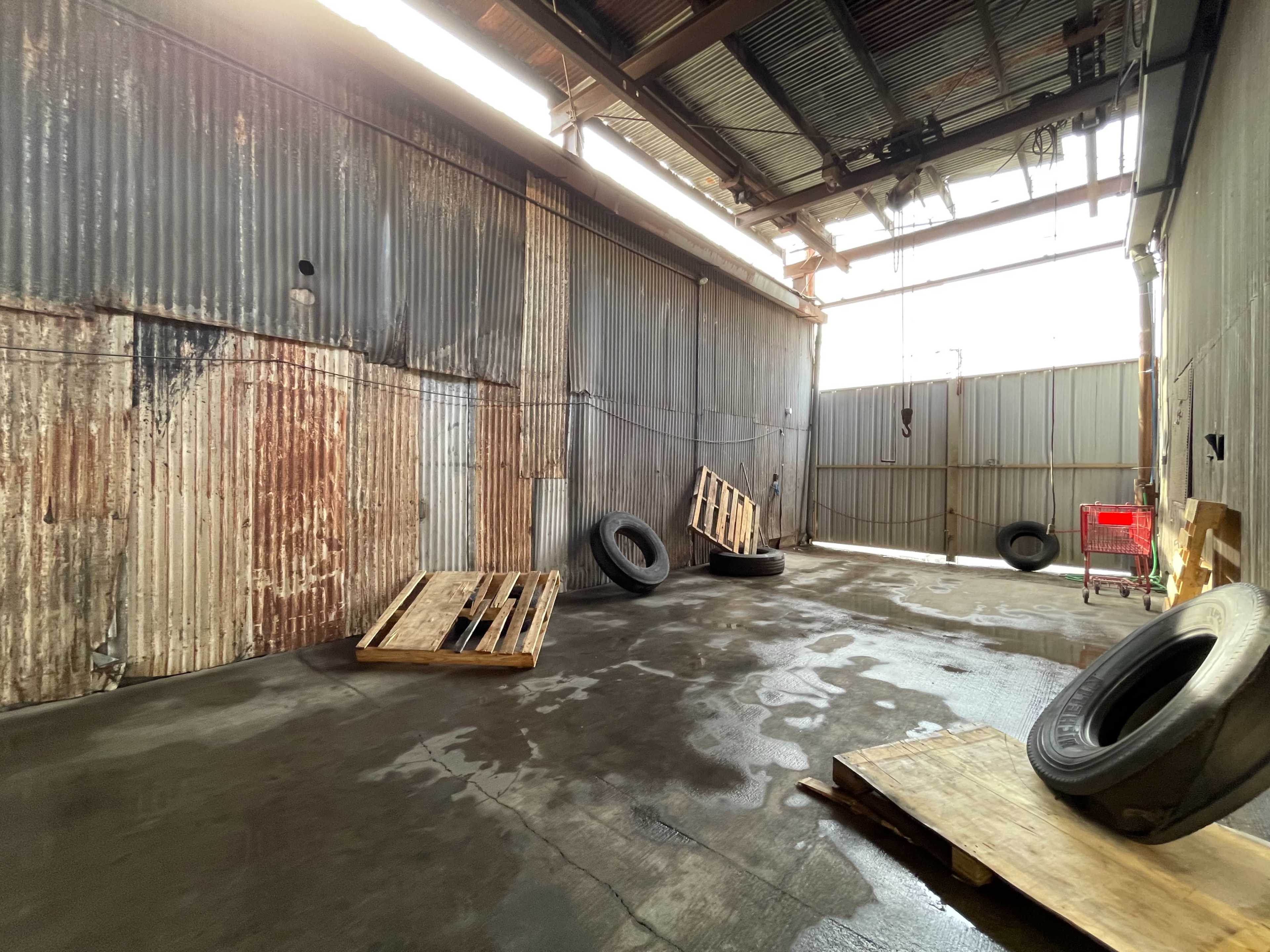An industrial space with a concrete floor, two leaning tires, wooden pallets, and a red shopping cart in a corner.