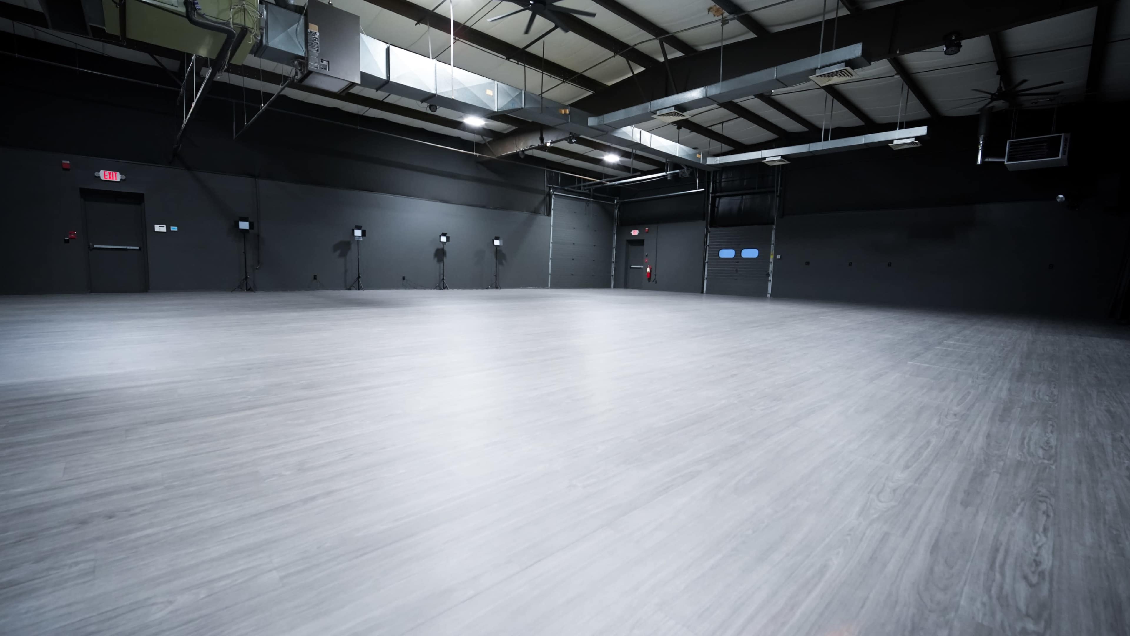 The image shows a spacious, empty interior with a gray wooden floor and black walls, featuring several overhead lights and large doors.