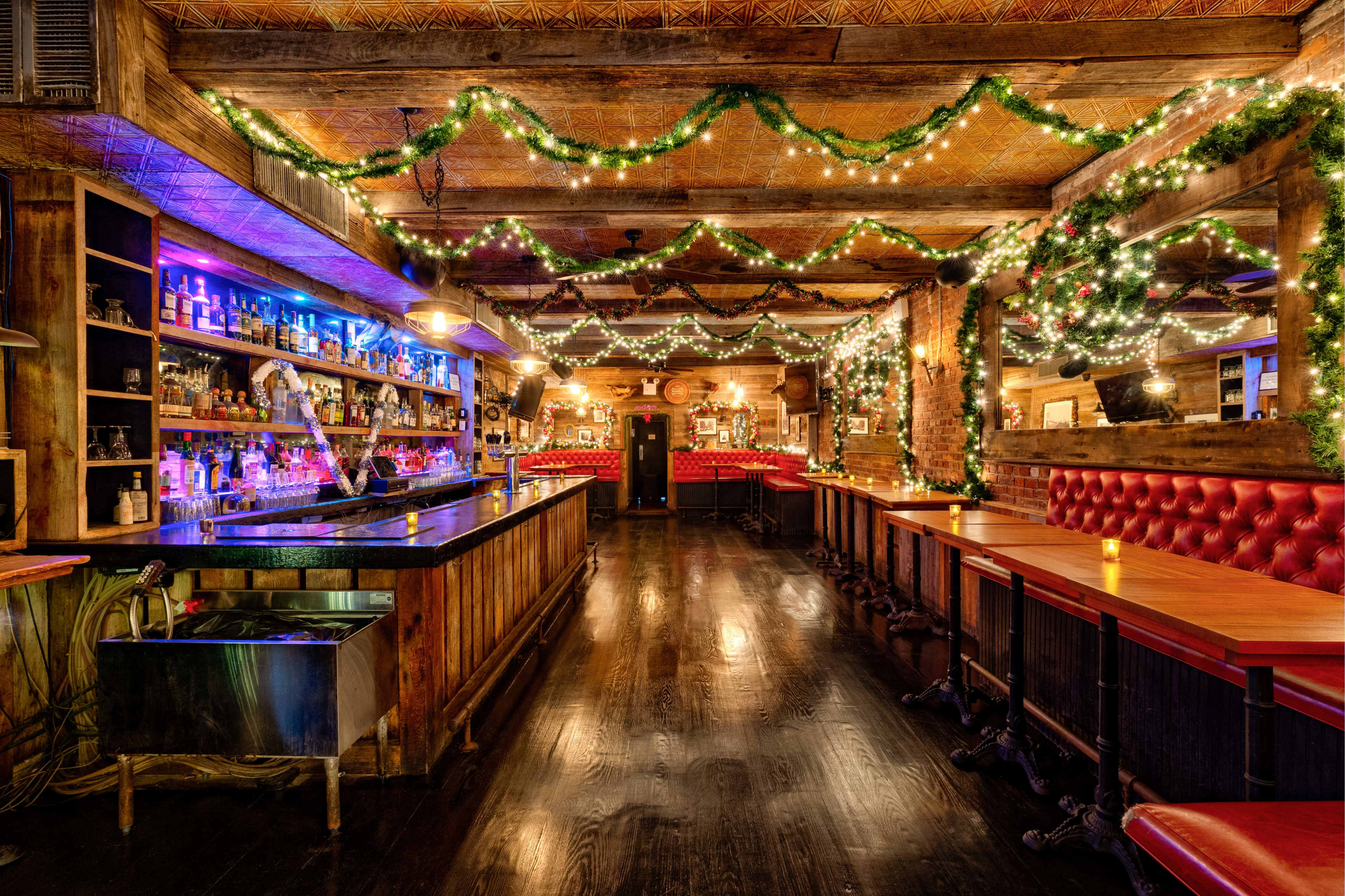 The image shows a cozy bar interior decorated with greenery and string lights, featuring a long bar and tables arranged along the sides.