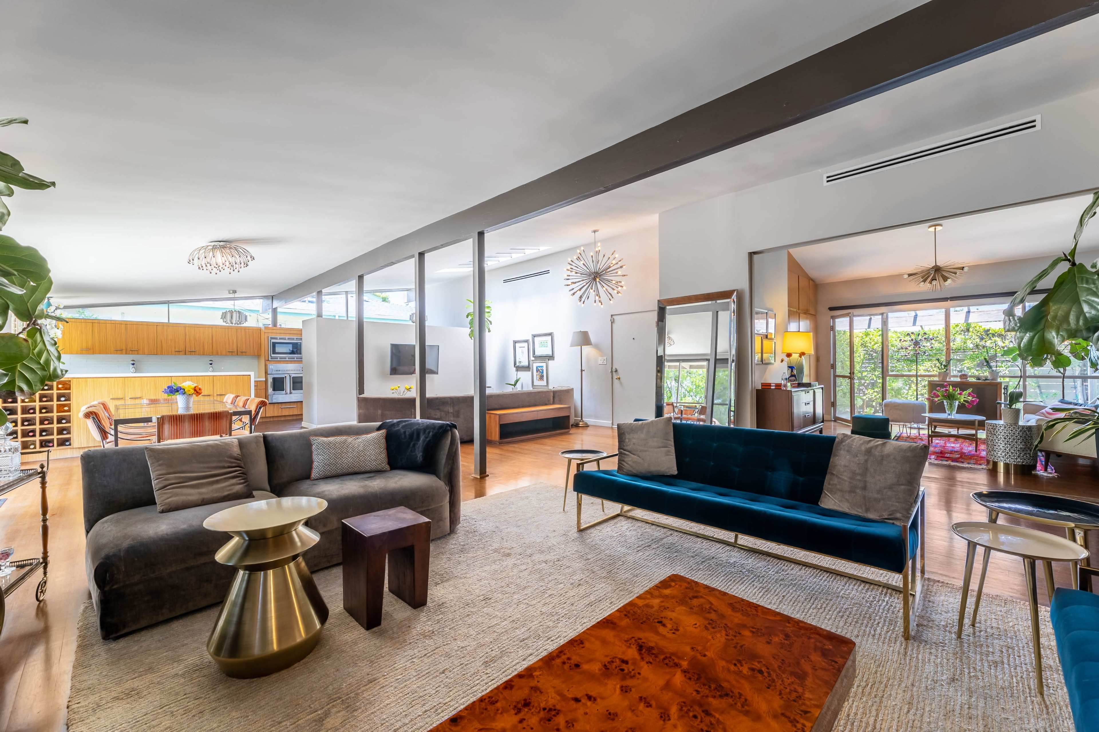 A spacious living area with modern furniture, including a dark sofa, blue velvet chairs, and a round coffee table, leading into a dining area with a wooden table and wine storage.