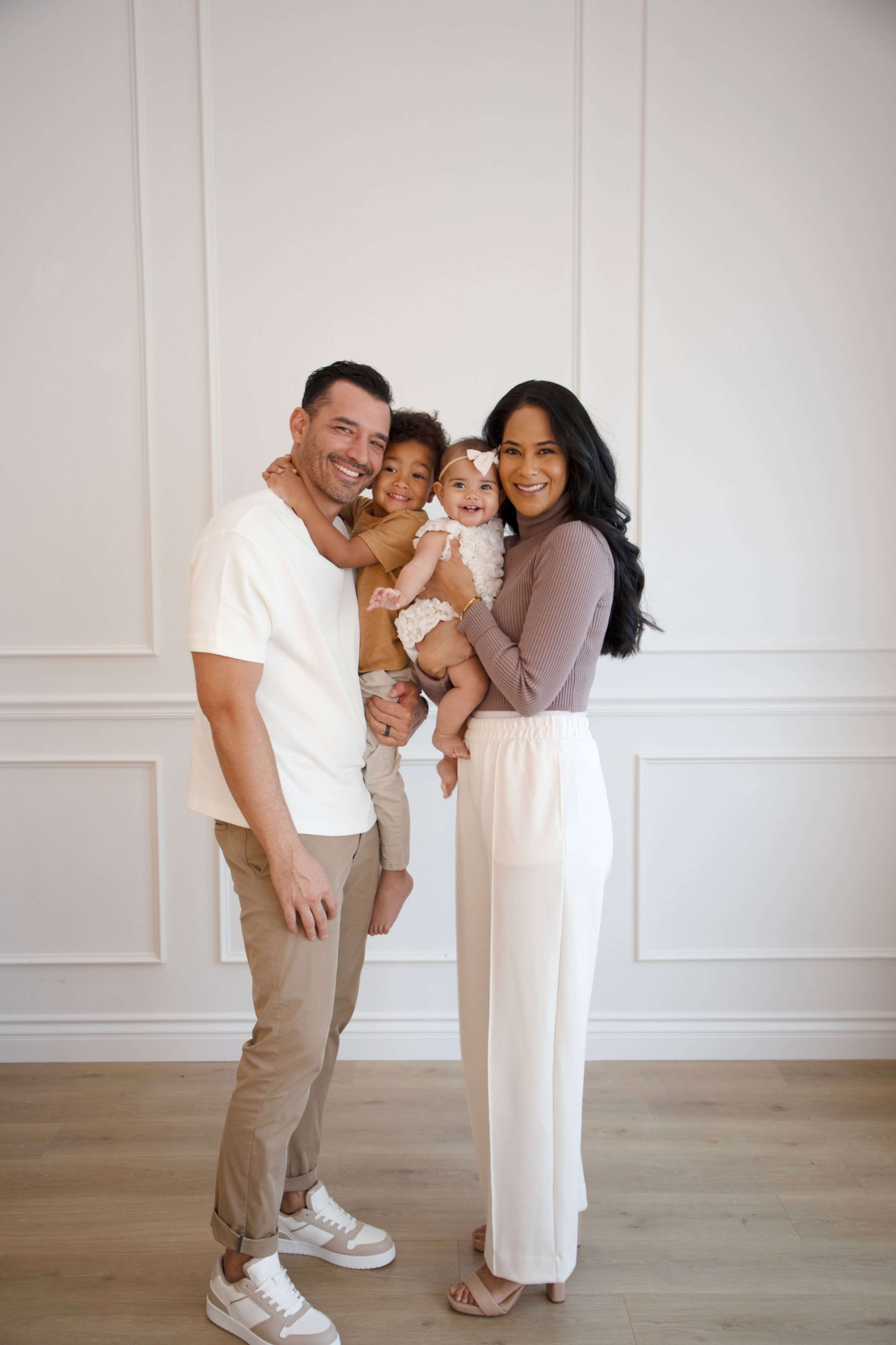 A couple stands together, smiling while holding two small children against a plain white wall.