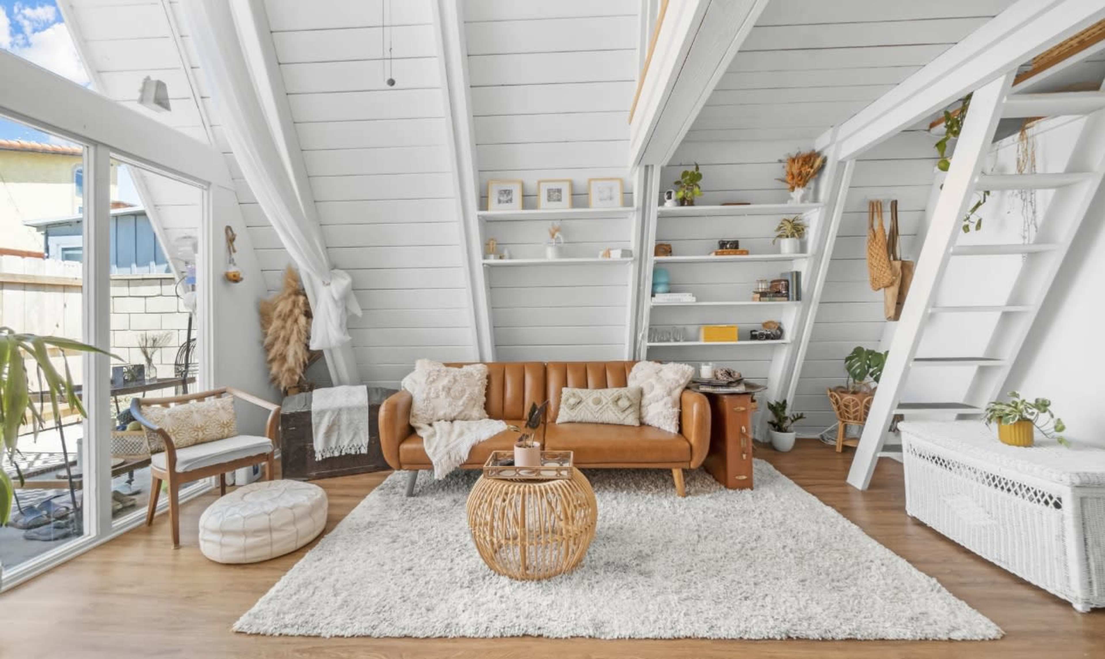 A cozy living room with a brown leather sofa, a round woven coffee table, and large windows surrounded by white walls and wooden accents.