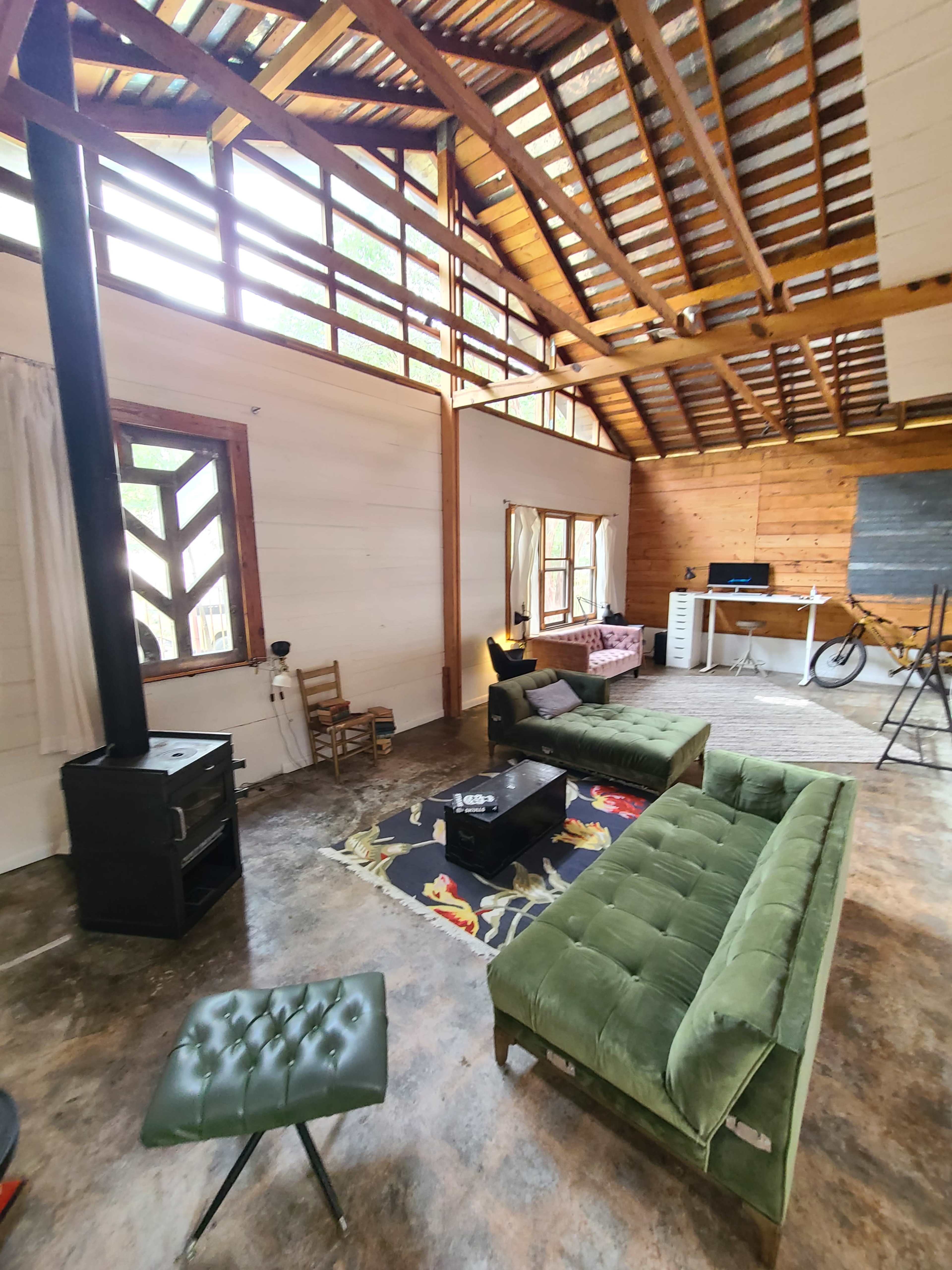 The image shows a spacious living area with exposed wooden beams, two green sofas, a black stove, a small table, and a workspace visible in the background.