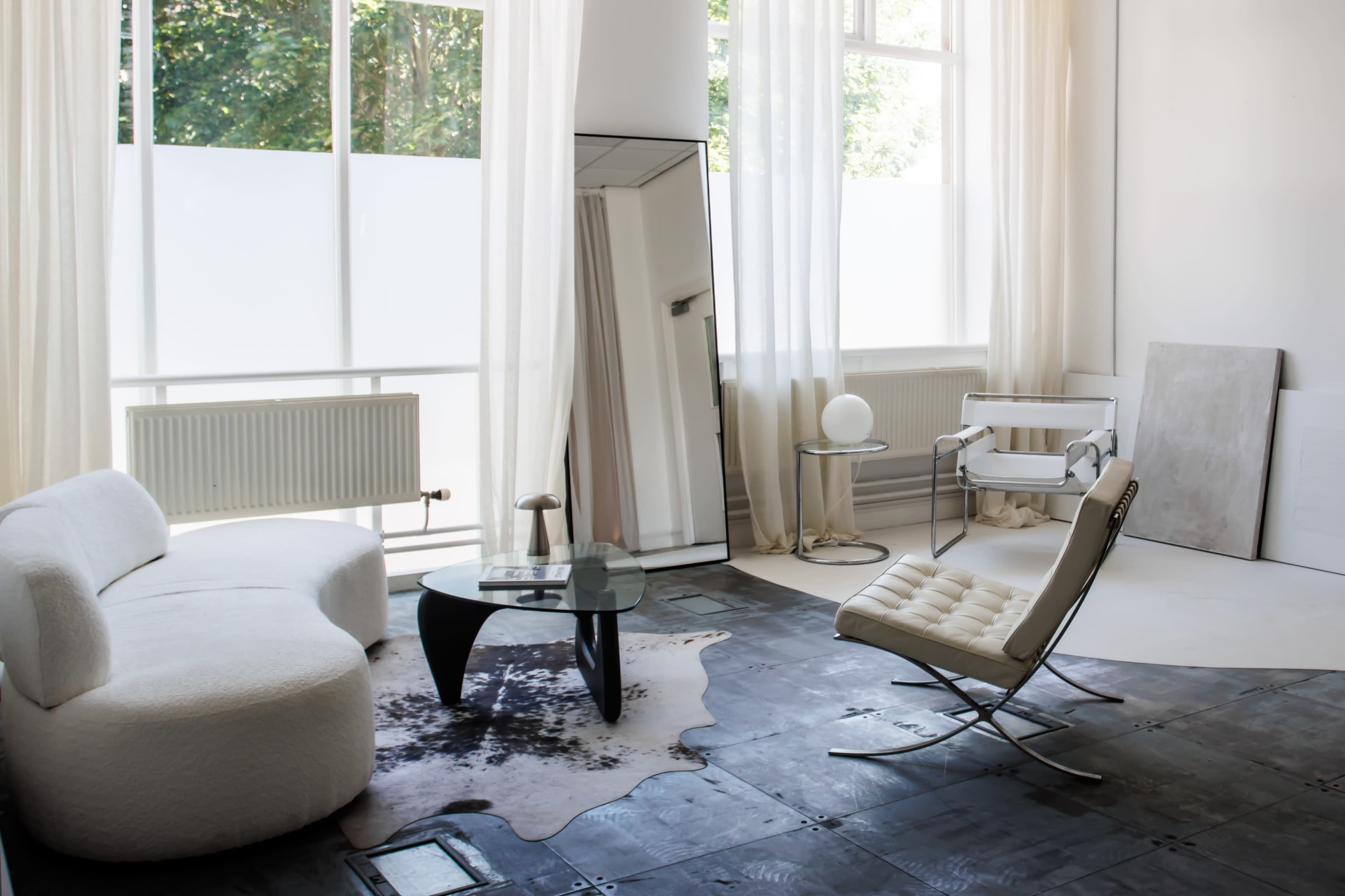 A bright, minimalistic living space features a white upholstered sofa, a glass coffee table, and a contemporary lounge chair, with large windows allowing ample natural light.