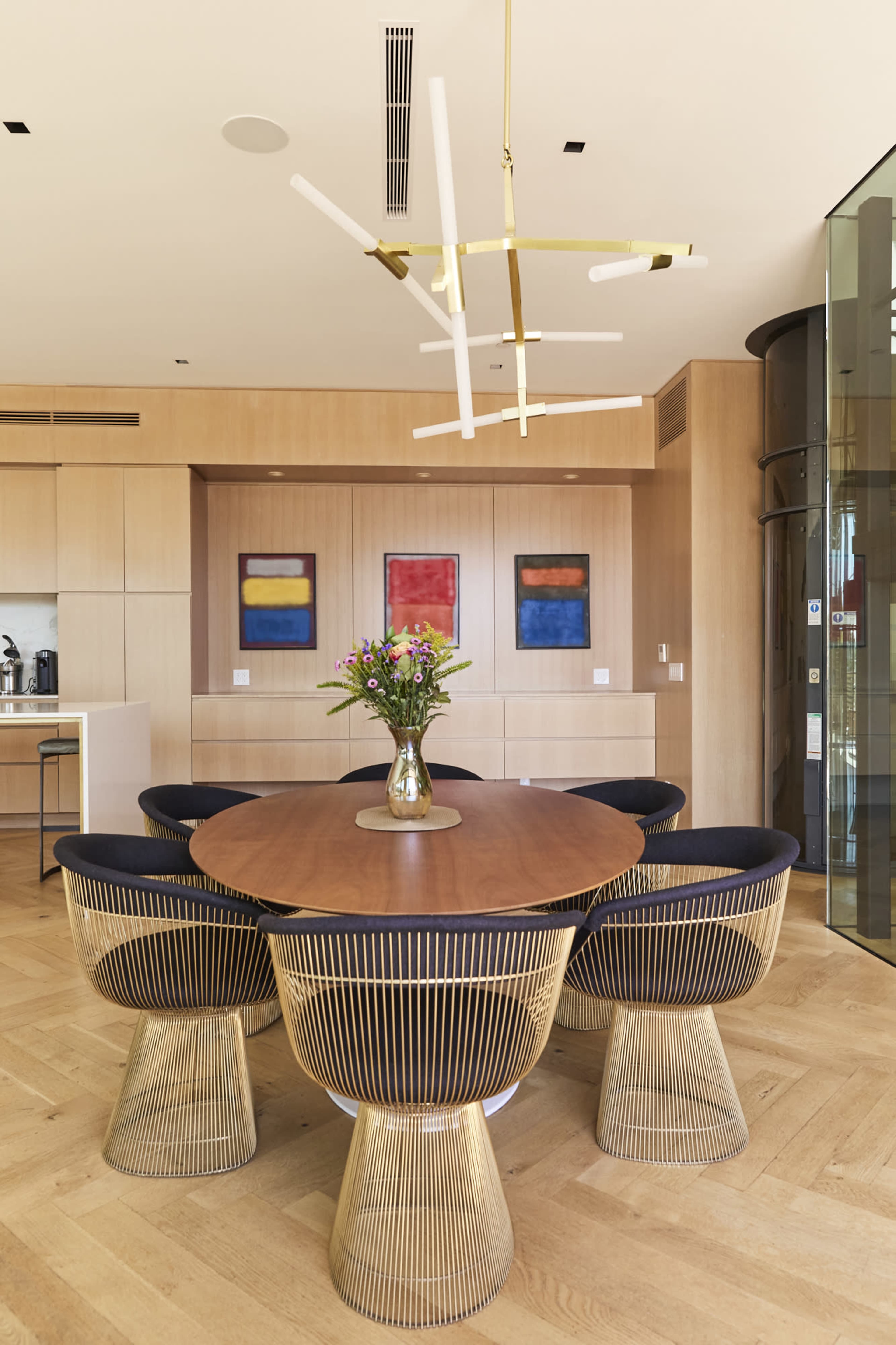 A round wooden dining table surrounded by six wireframe chairs is set in a modern dining area featuring a floral centerpiece and abstract paintings on the walls.