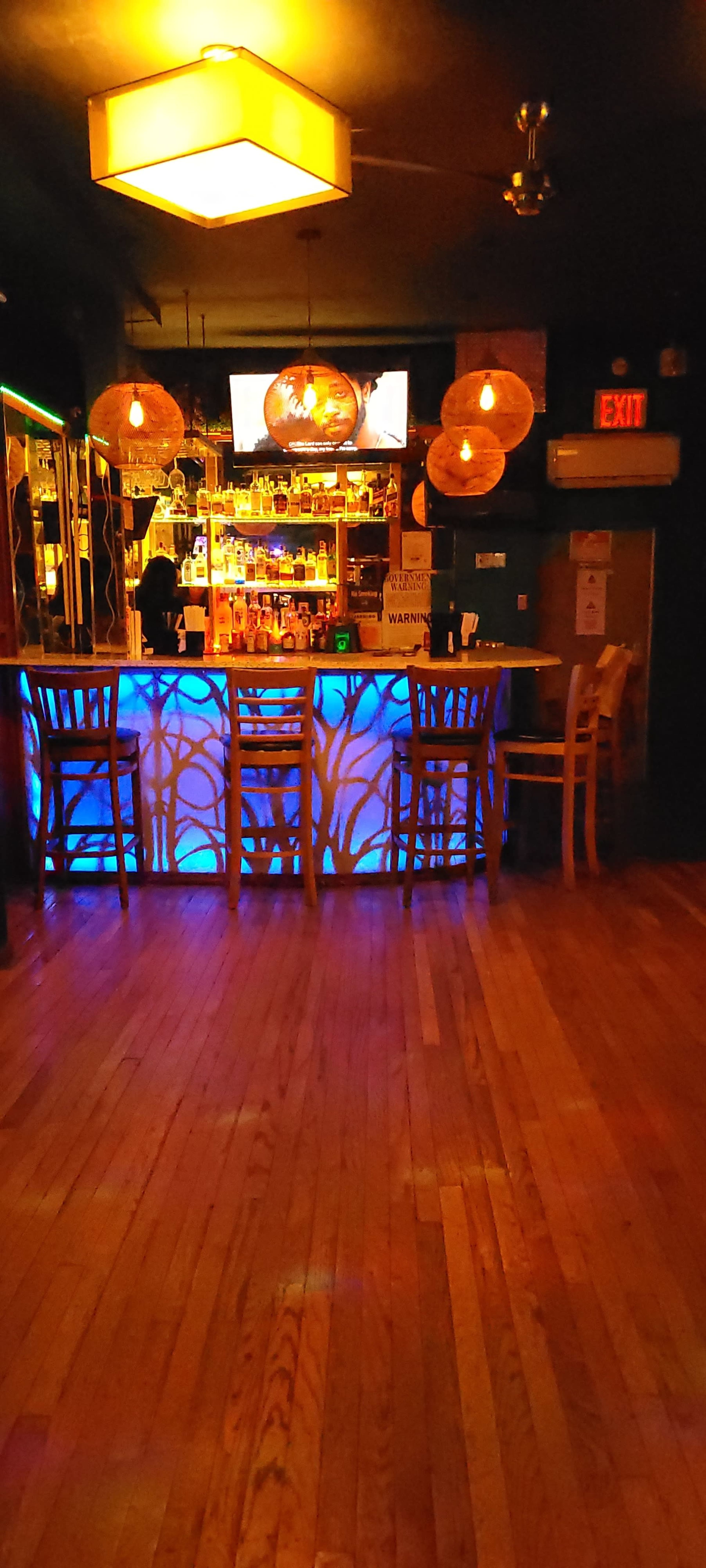 The image shows a dimly lit bar area featuring wooden flooring, a glowing blue bar, and several hanging pendant lights.