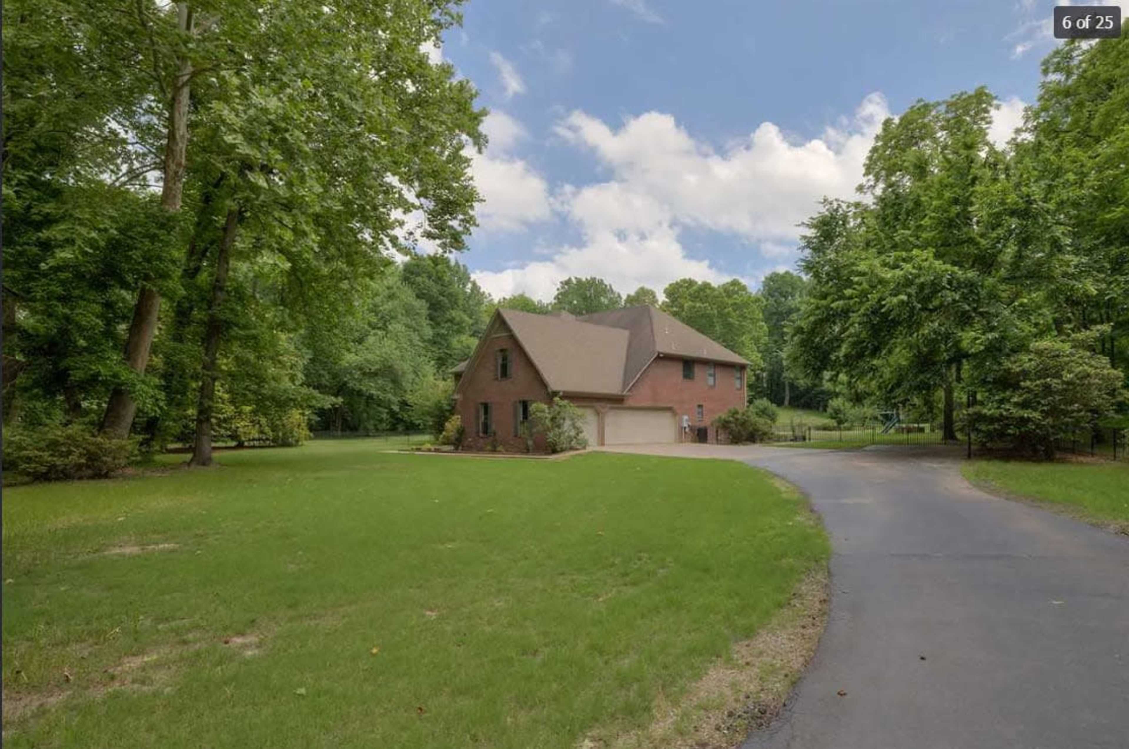 A brick house is set back from a winding driveway, surrounded by lush green trees and a large open lawn.