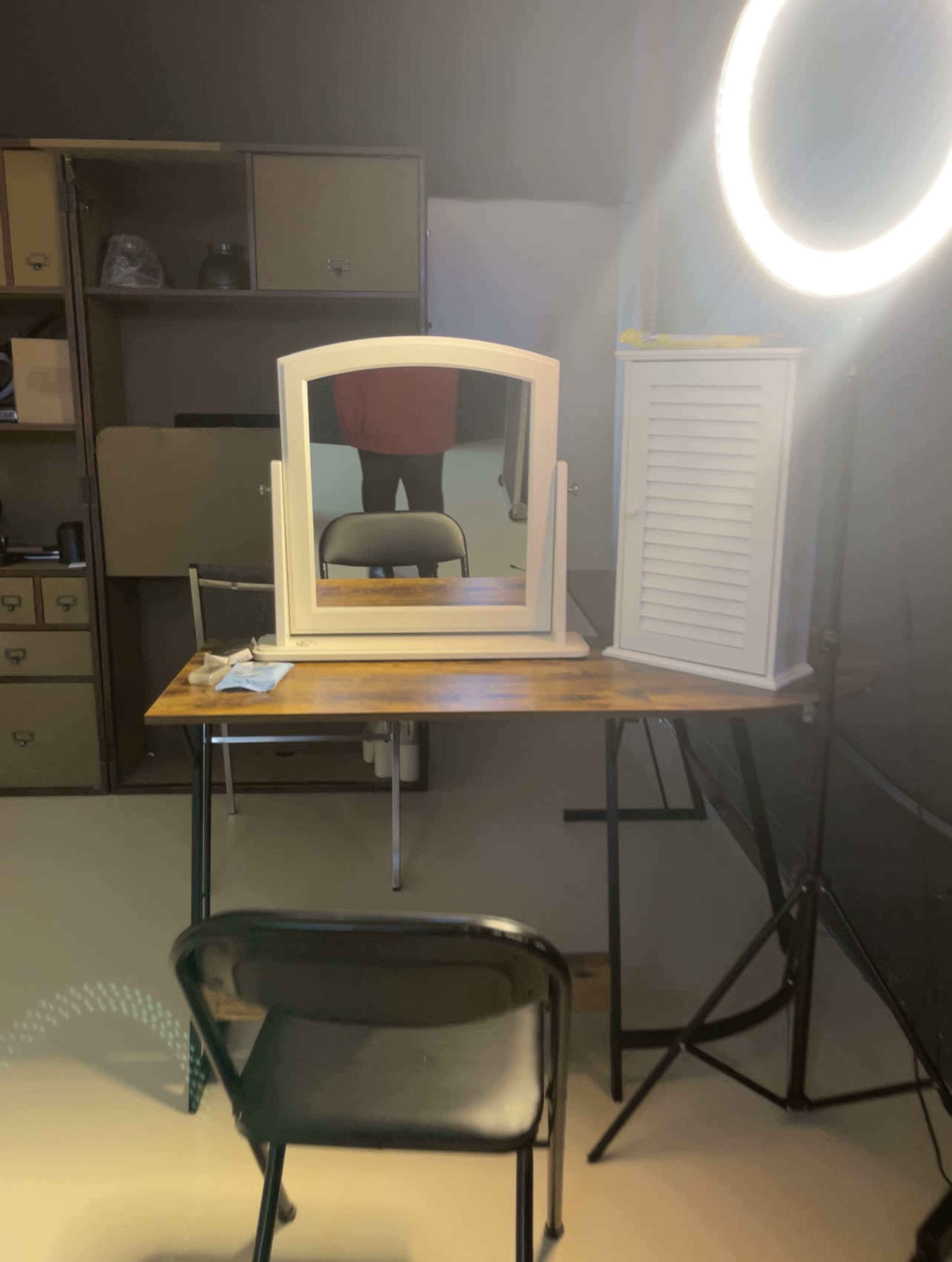 A wooden table with a white-framed mirror and a folding chair is positioned in front of a ring light in a room with storage cabinets.
