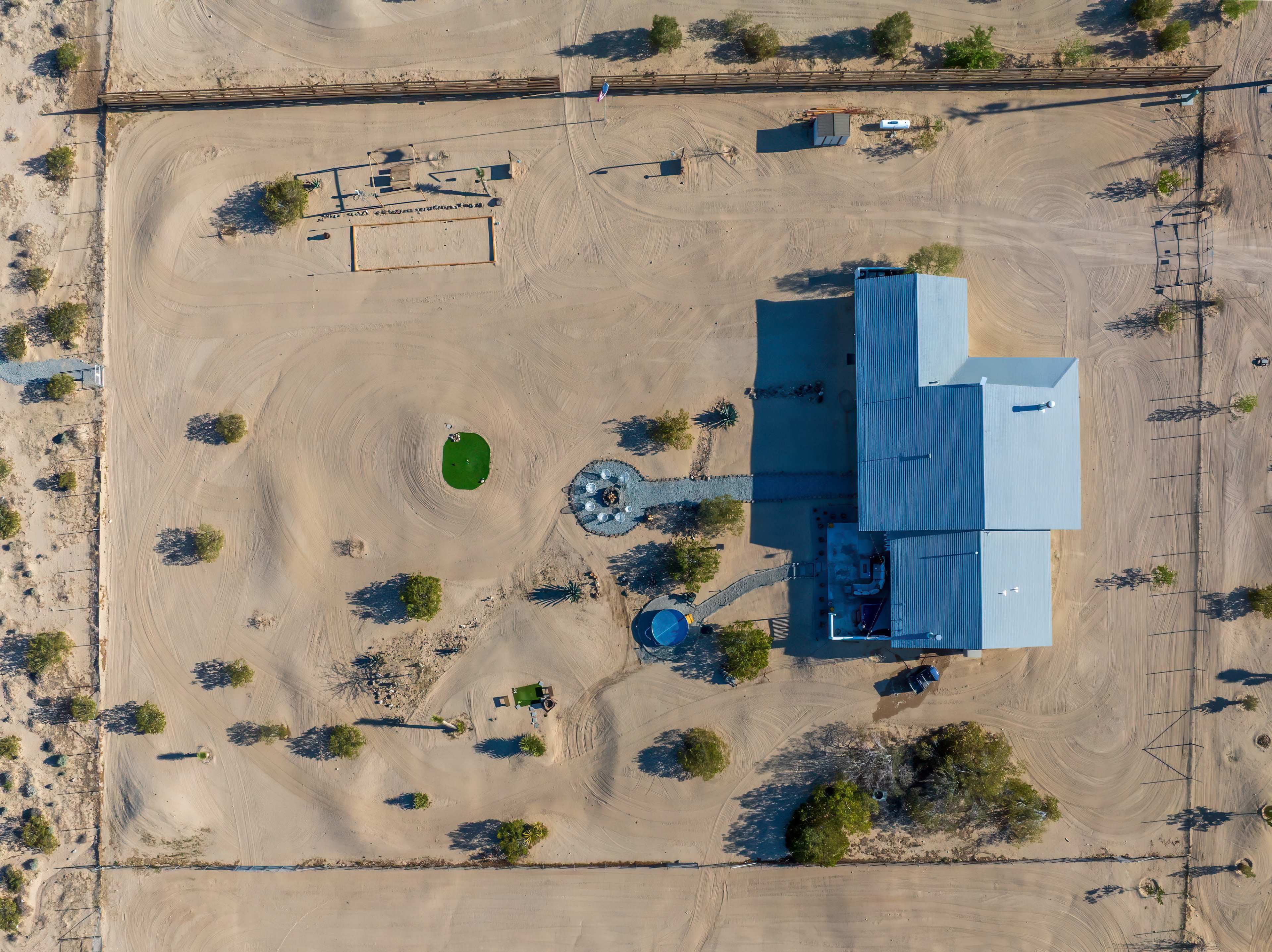 The image shows an aerial view of a large building surrounded by a sandy landscape, with sparse vegetation and various outdoor fixtures.