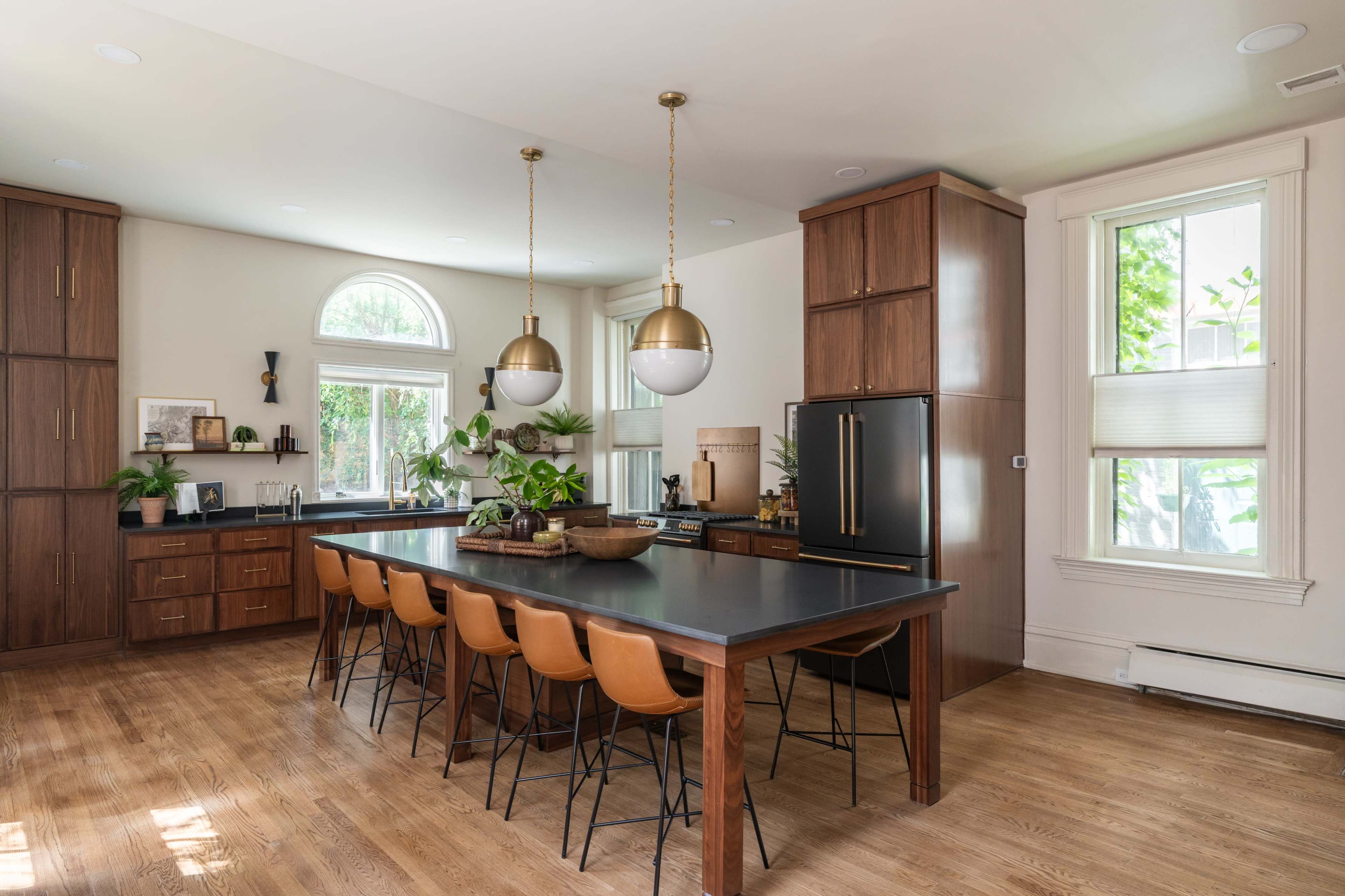 The image shows a modern kitchen with a large island featuring brown leather seating, wooden cabinetry, and large windows allowing natural light.