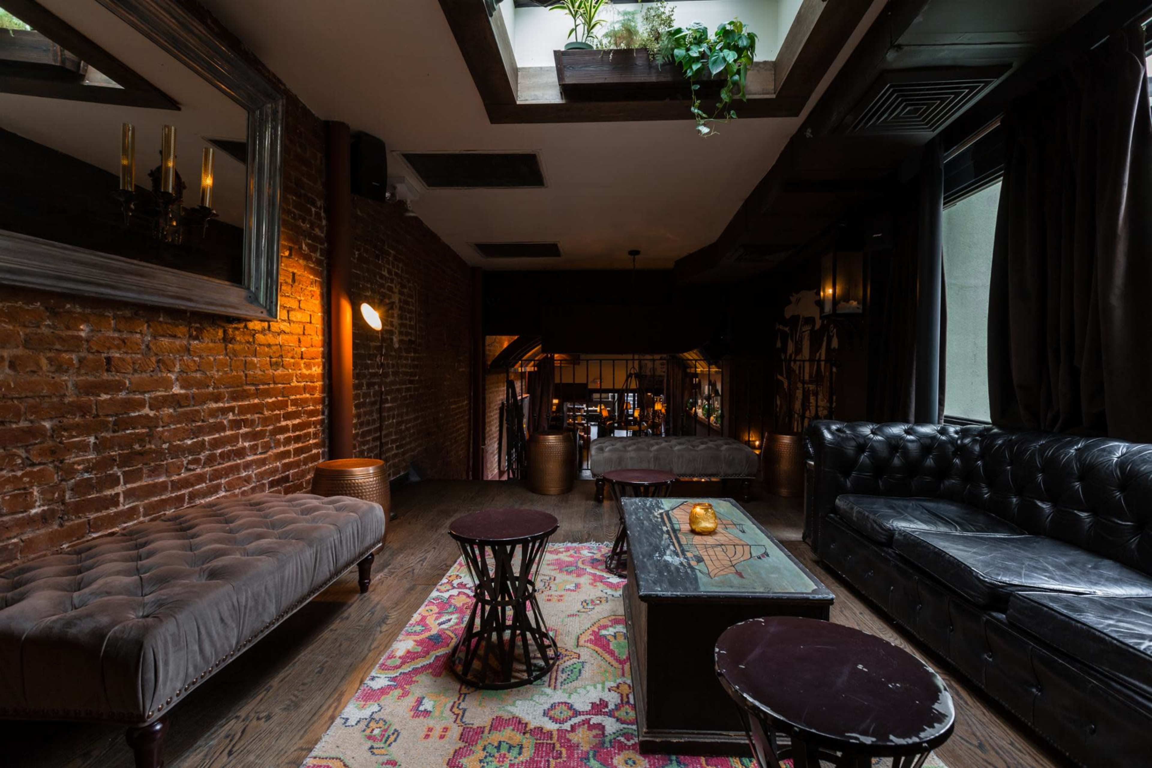 A dimly lit lounge with exposed brick walls, a tufted velvet sofa, a wooden coffee table, and patterned rugs.