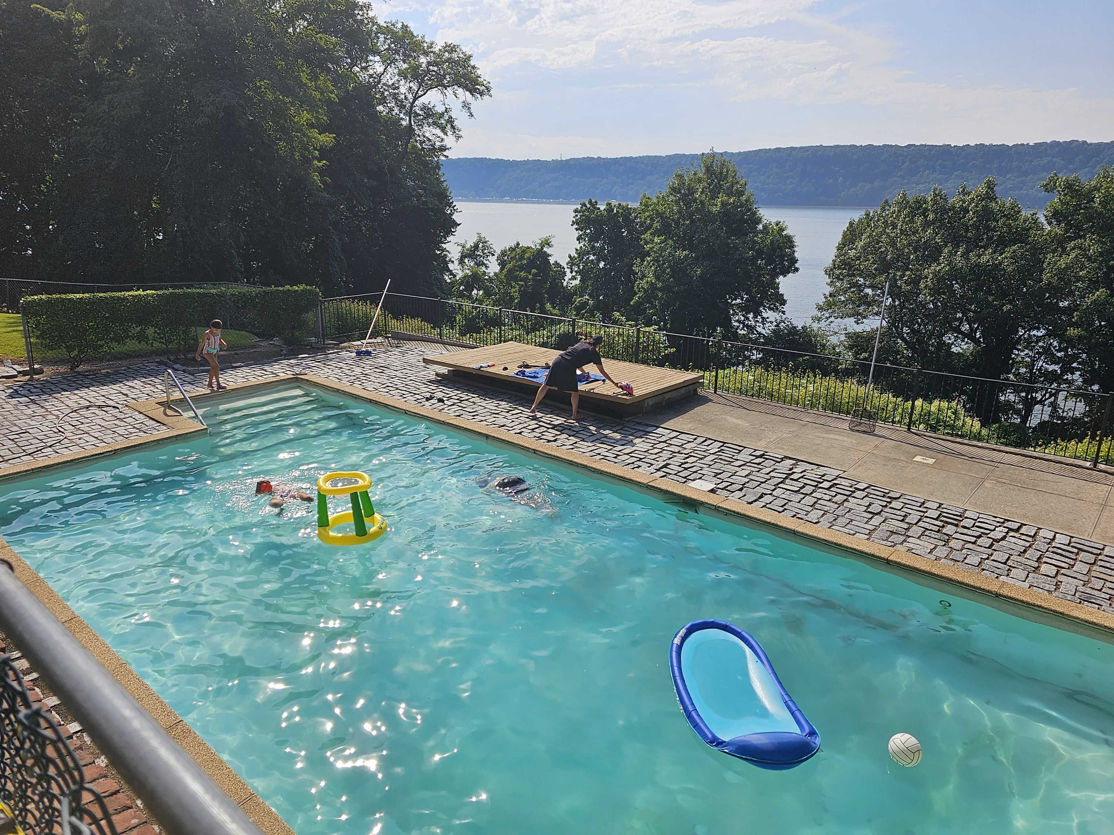 The scene shows a swimming pool surrounded by green foliage, with two children playing in the water and an adult stretching on the pool's edge, while a scenic riverbank is visible in the background.