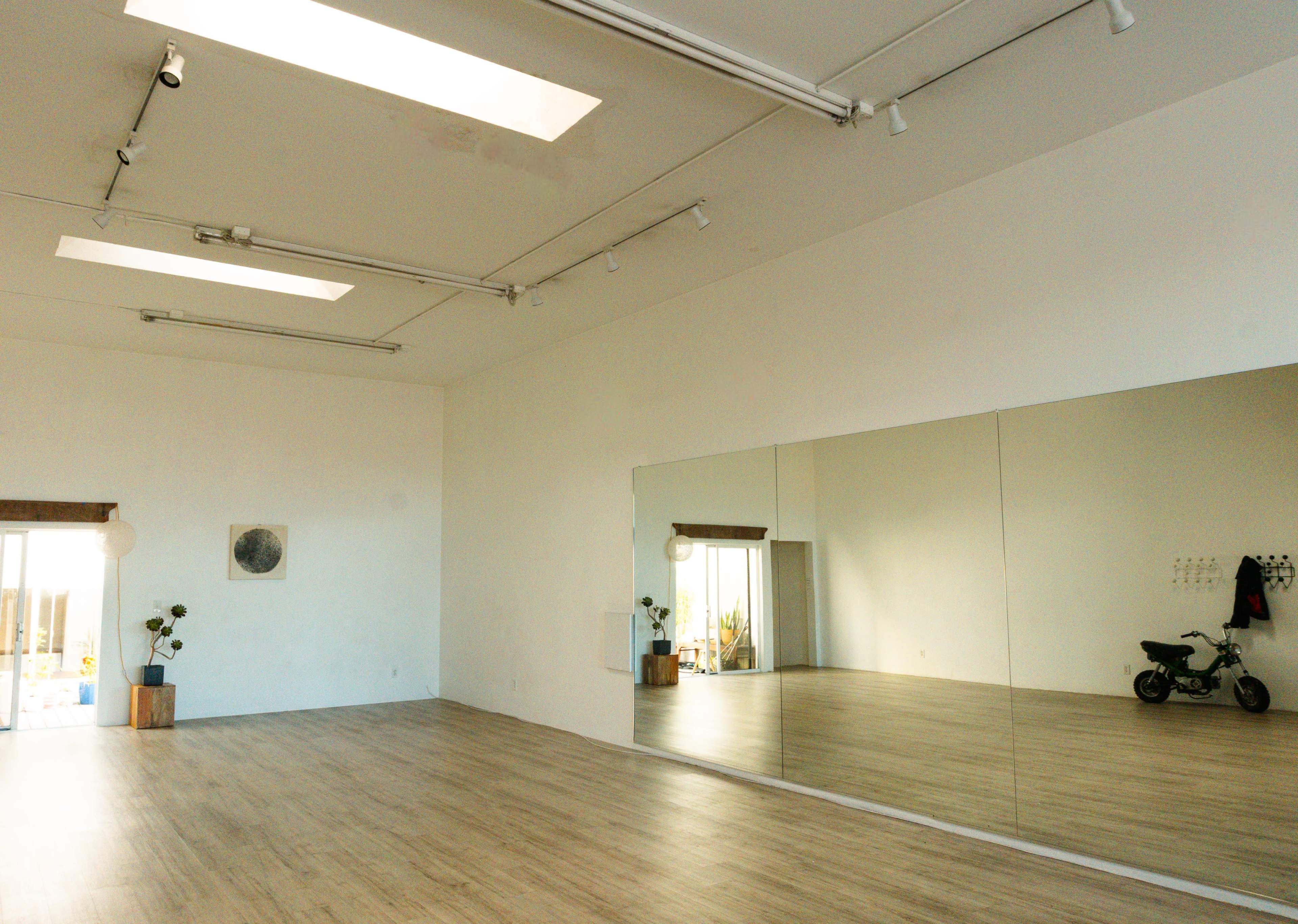 The image shows a spacious room with wooden flooring, large mirrors on one wall, and natural light streaming through skylights and a window.