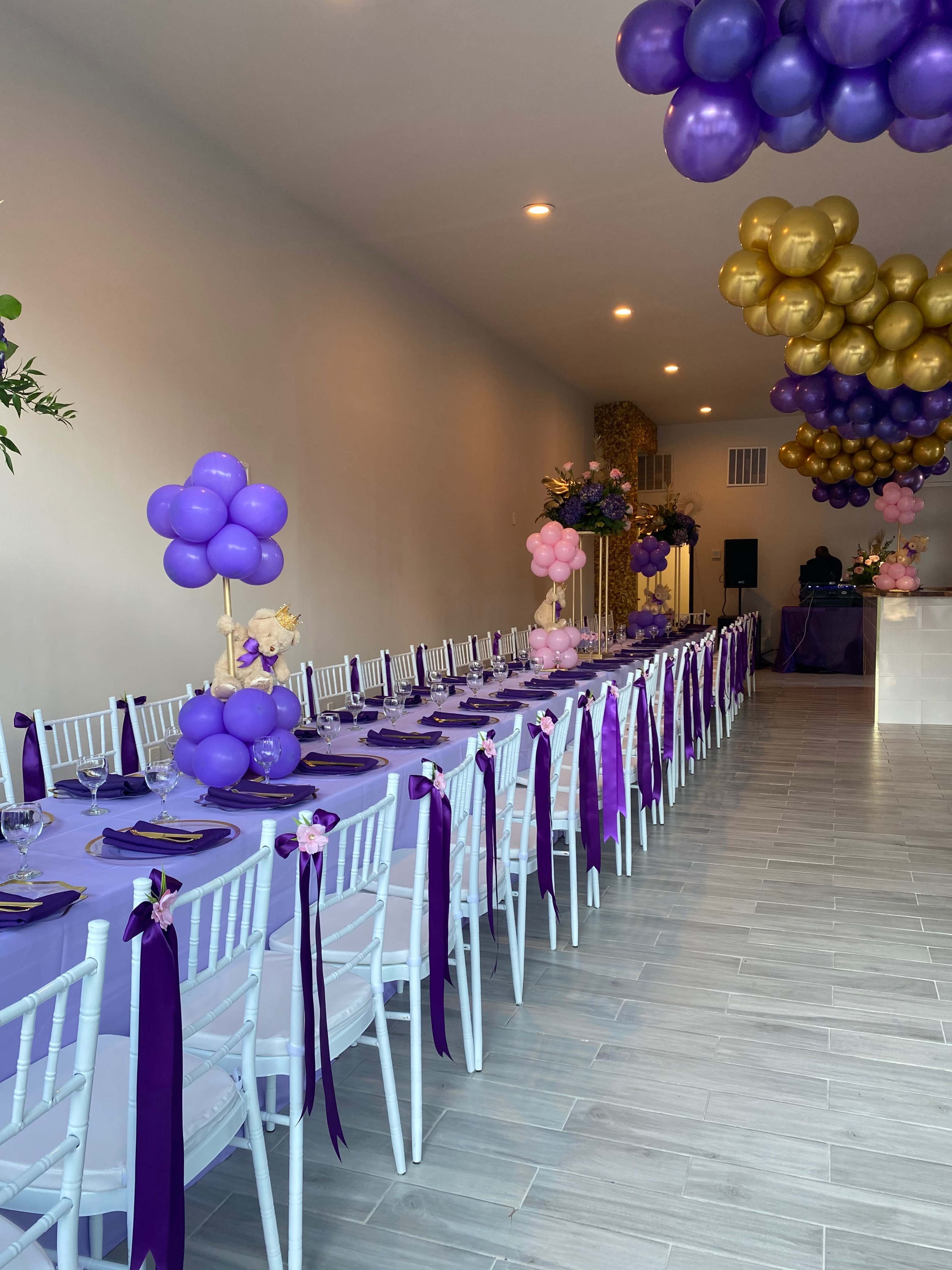 A long table with purple linens and white chairs, decorated with balloon centerpieces and set for a celebration.
