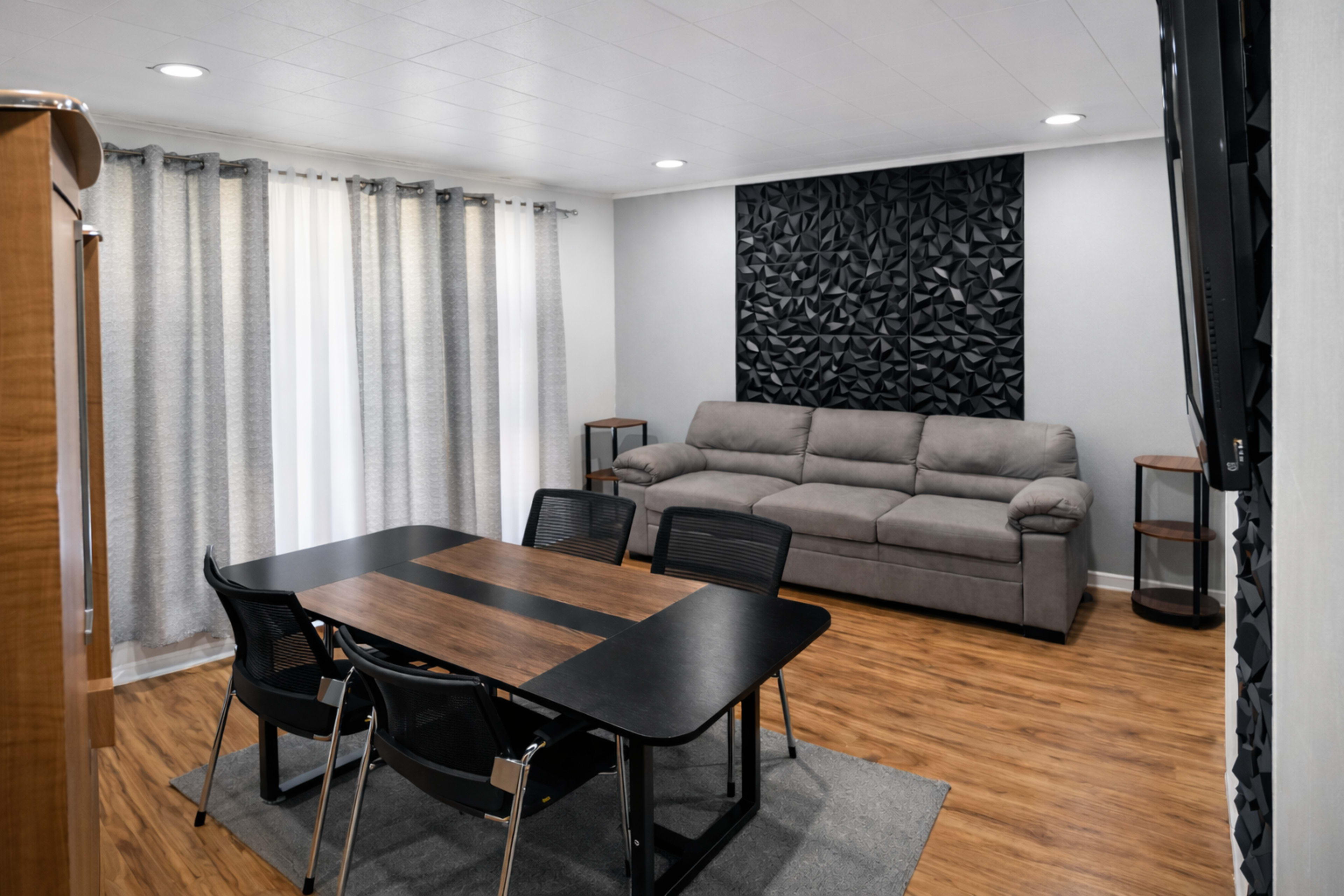 The image shows a modern living space featuring a dining table with four black chairs, a gray sectional sofa, and a textured black wall panel, illuminated by natural light from the windows.