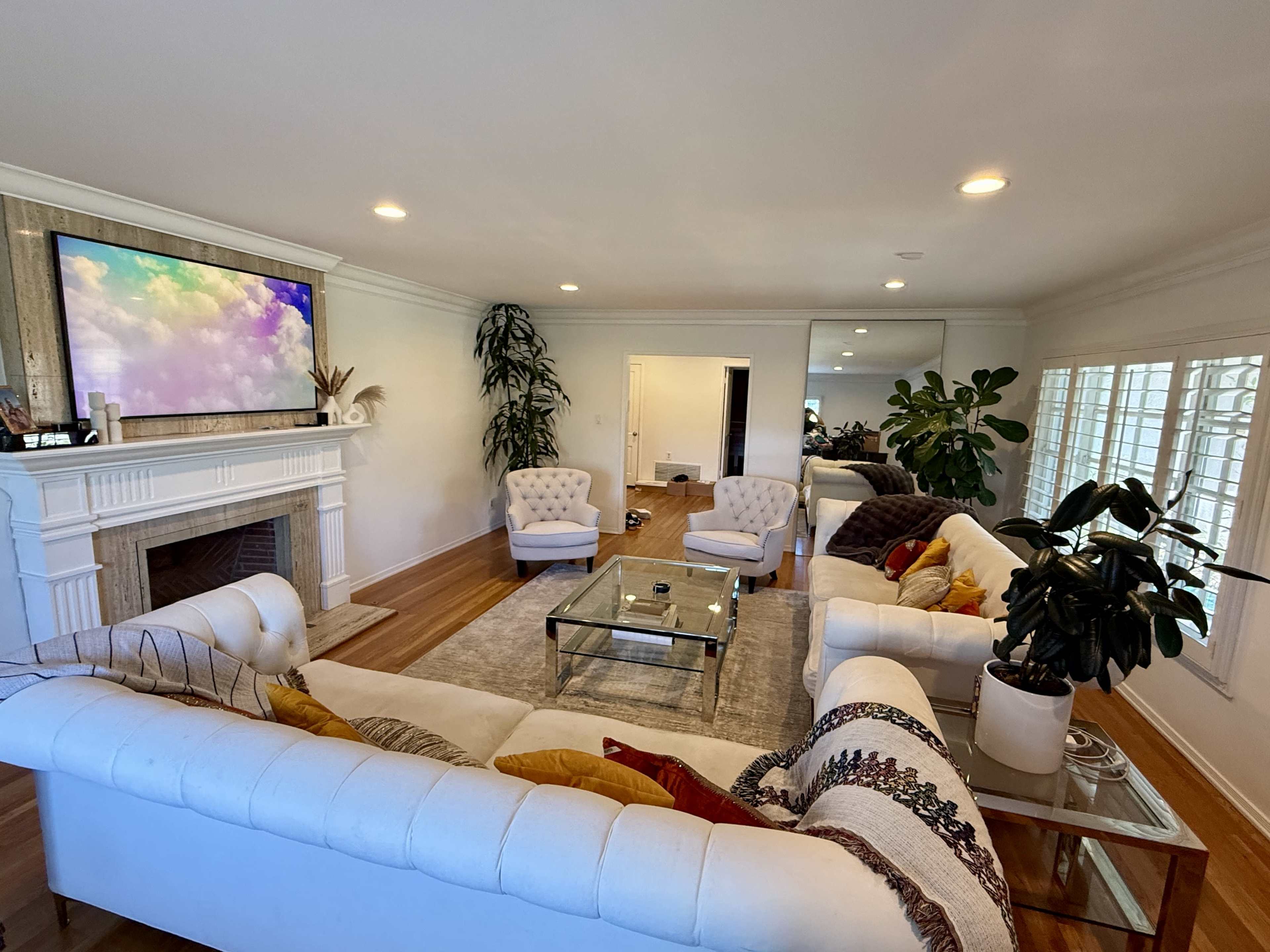 The living room features two white upholstered sofas, a glass coffee table, and a large TV mounted above a fireplace, with indoor plants in the corners.