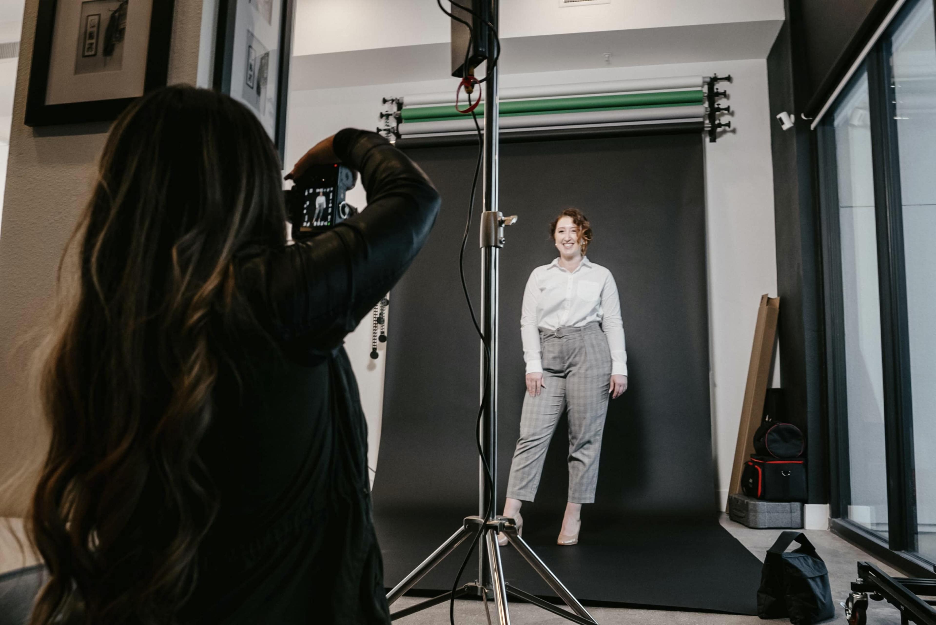 A woman stands posing in front of a camera on a photography set with a dark backdrop.
