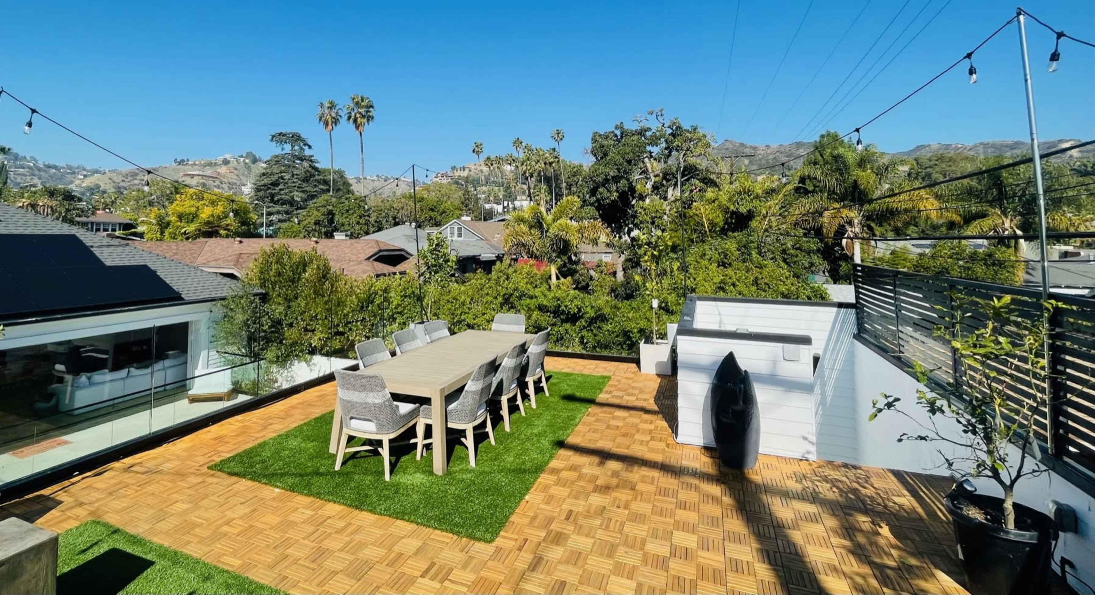 A rooftop terrace featuring a large dining table surrounded by chairs, artificial grass, and views of palm trees and distant hills.