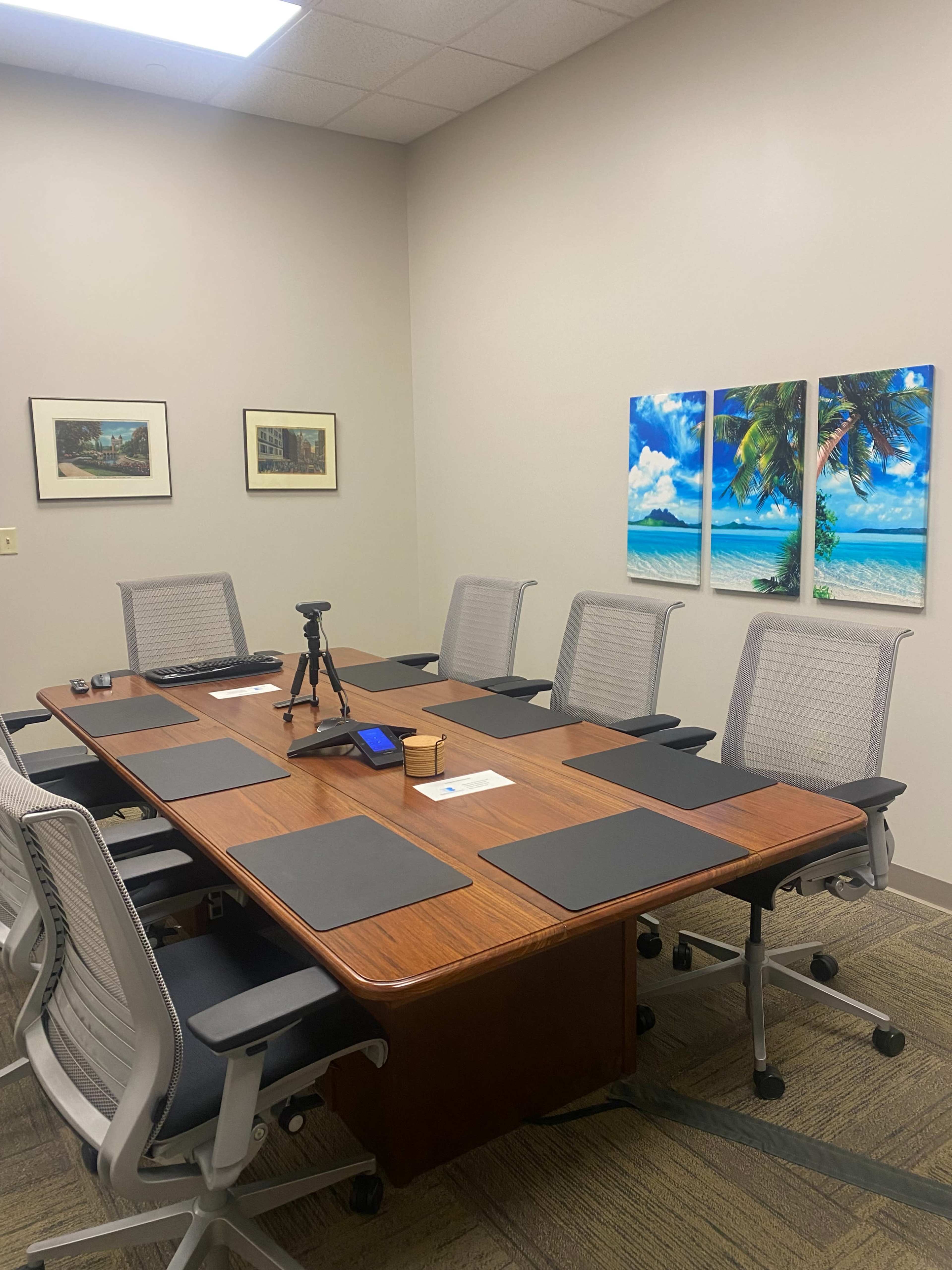 A conference room with a large wooden table surrounded by eight chairs, equipped with a tripod and various office supplies, and decorated with two palm tree paintings on the wall.
