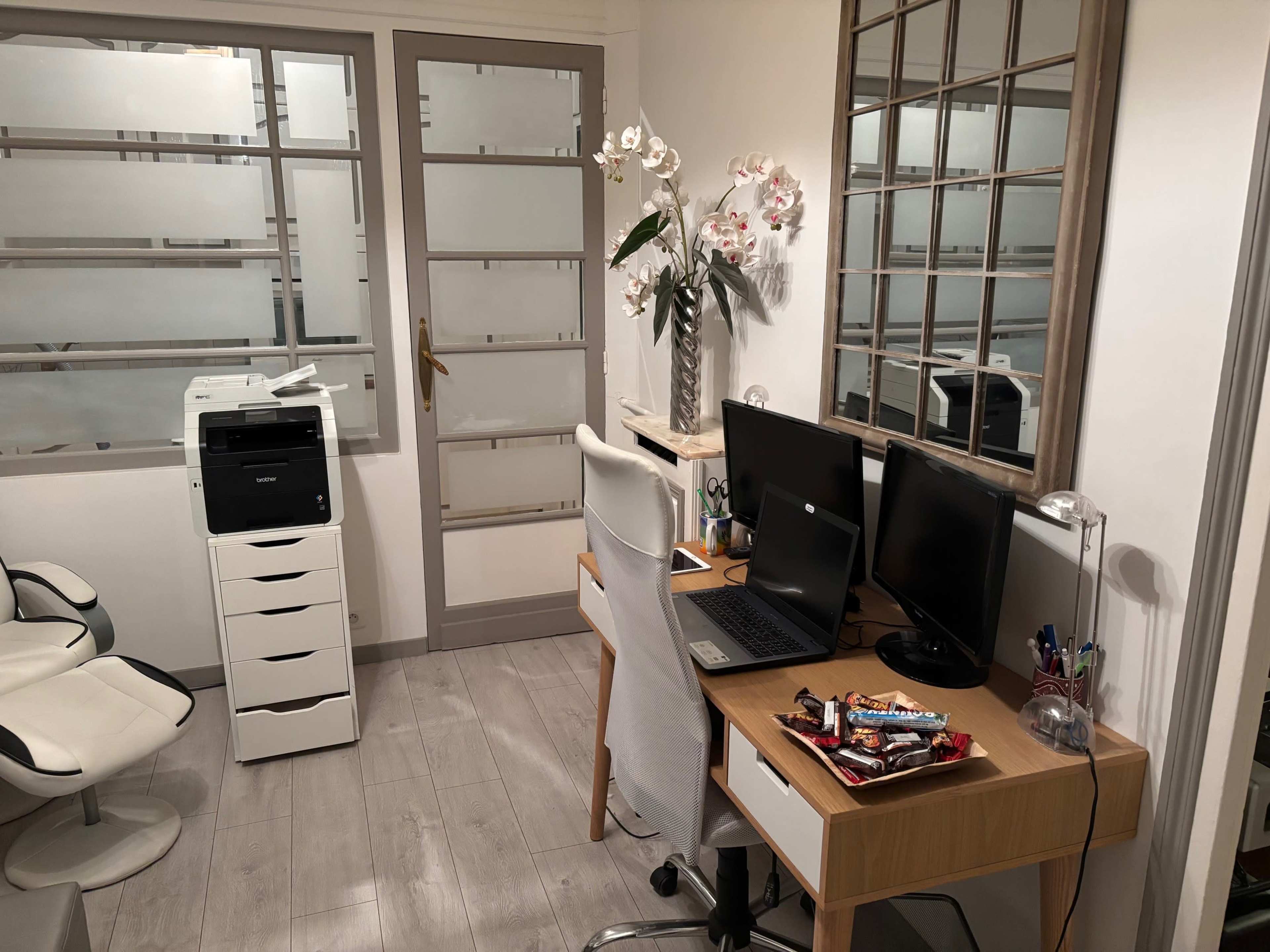 The image shows an office space with a desk featuring two computer monitors, a printer, and a vase with flowers, alongside a storage unit and a chair positioned against the wall.