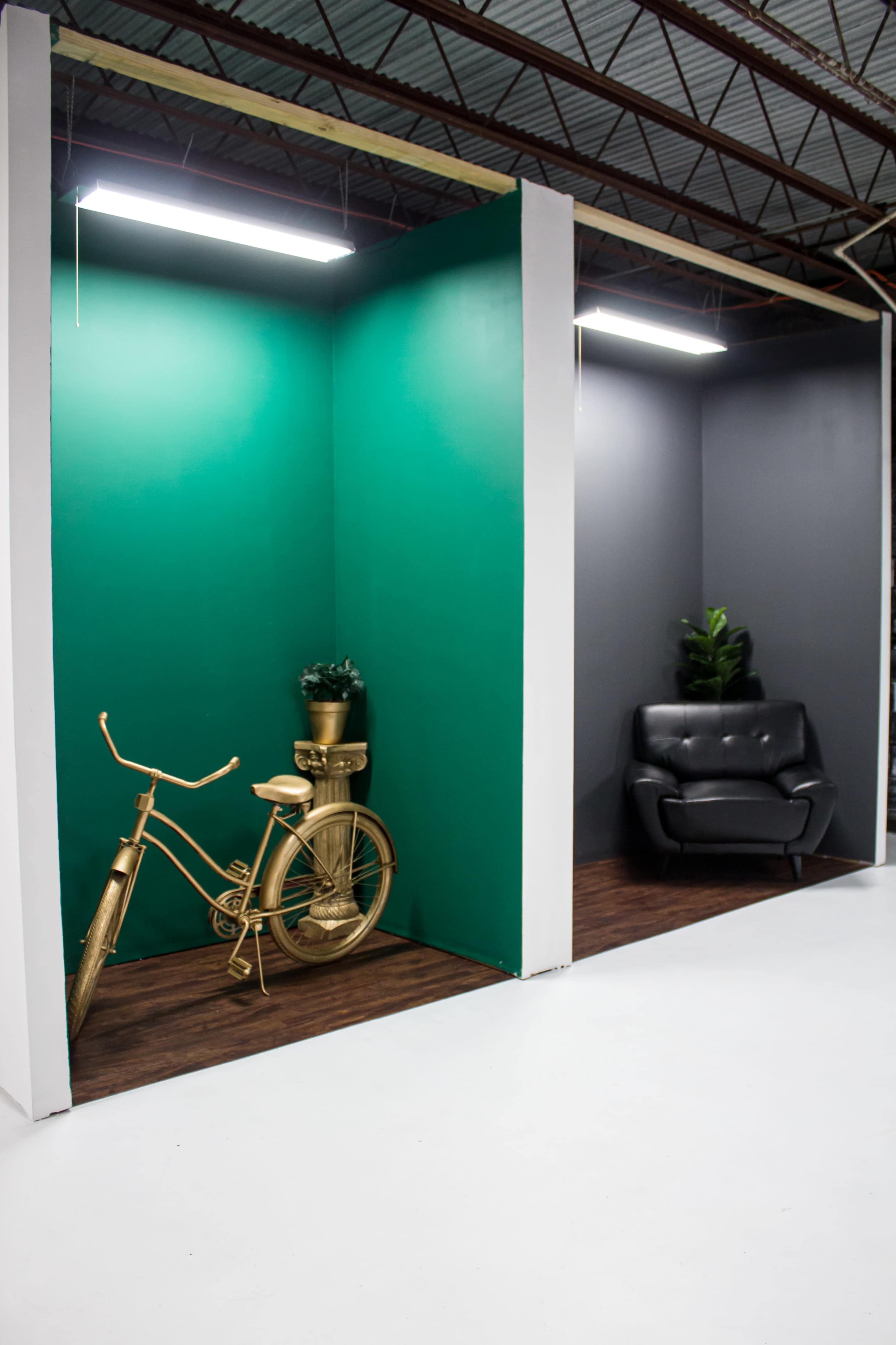 The image shows a divided interior space featuring a golden bicycle and a plant on one side, and a black chair surrounded by greenery on the other.